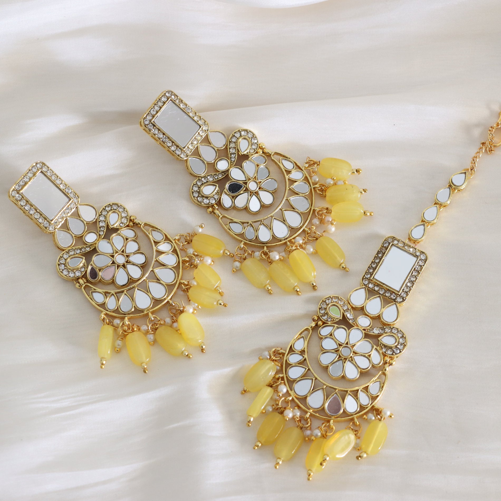 Gold-toned traditional Indian jewelry set featuring a matching dangle choker necklace and earrings with white inlay stones and yellow bead drops, arranged on a white satin fabric background.