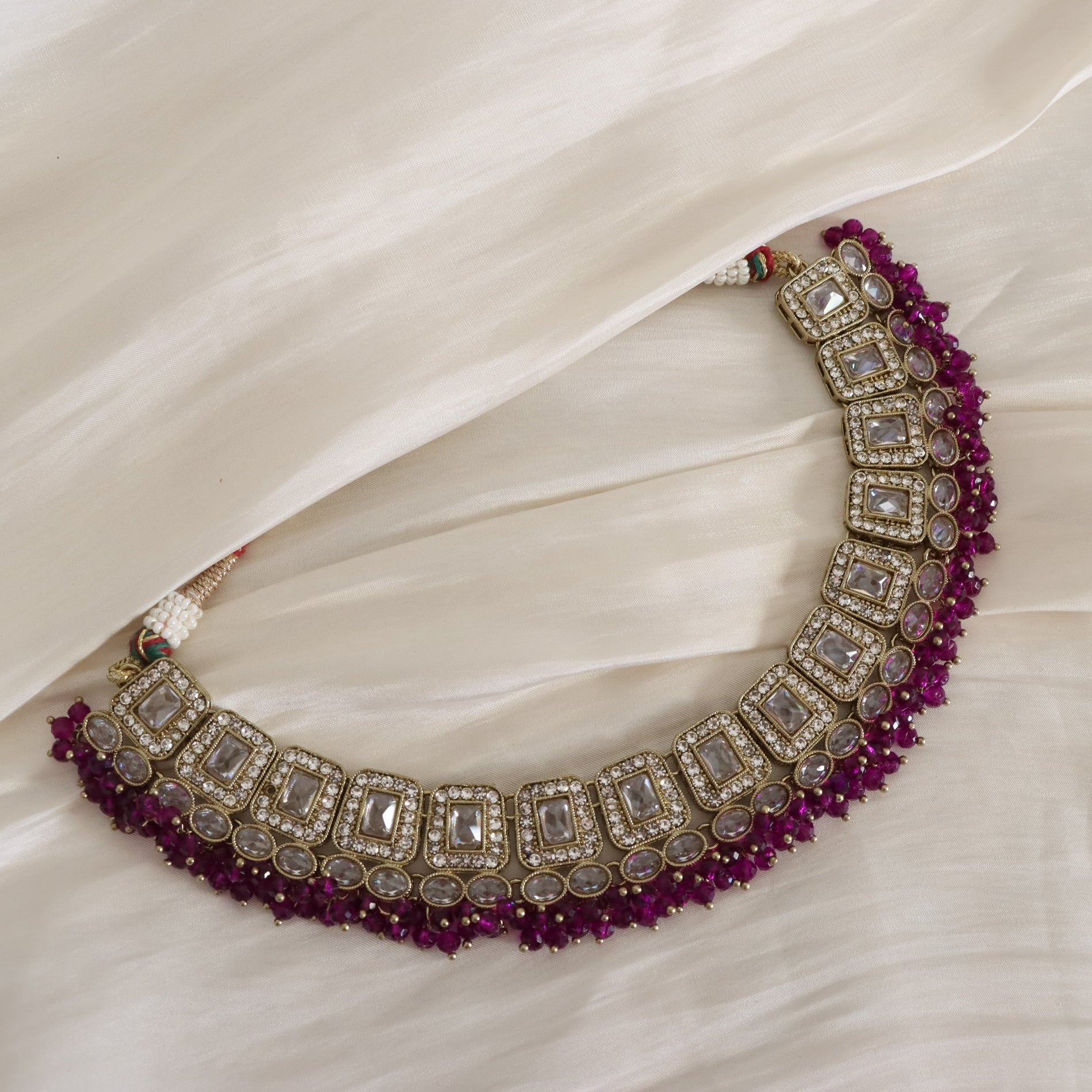 An ornate Indian-style choker necklace with silver-toned metal, clear stones, and deep magenta-pink beaded borders, displayed on a draped cream-colored fabric.