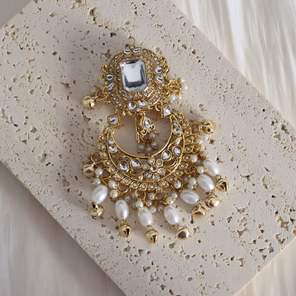 An ornate gold-toned statement earring decorated with clear crystals, white faux pearls, and small gold bells, displayed on a textured beige stone tile.
