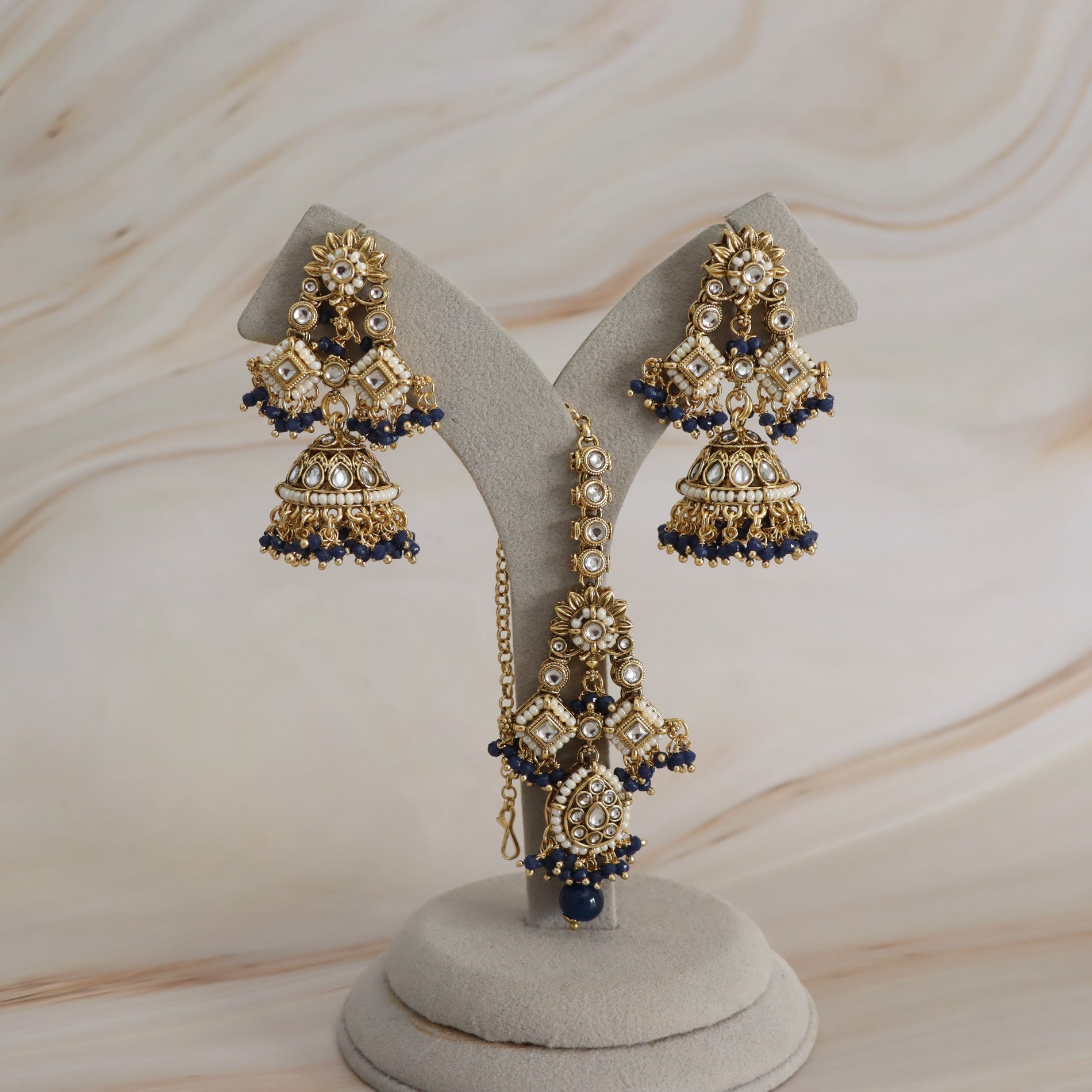 Gold-plated Indian jhumka earrings with white stones and dark blue bead accents, displayed on a gray velvet jewelry stand against a light wood background.