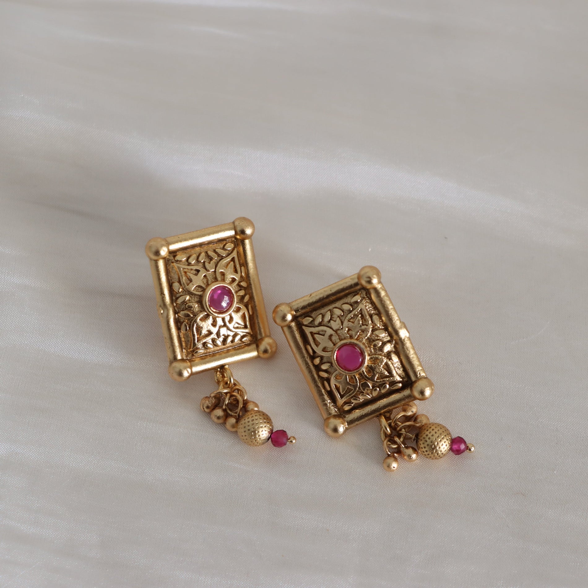 A pair of gold-toned rectangular earrings with intricate filigree detailing, a small pink gemstone in the center, and dangling gold beads and small pink accents, displayed on a light textured surface.