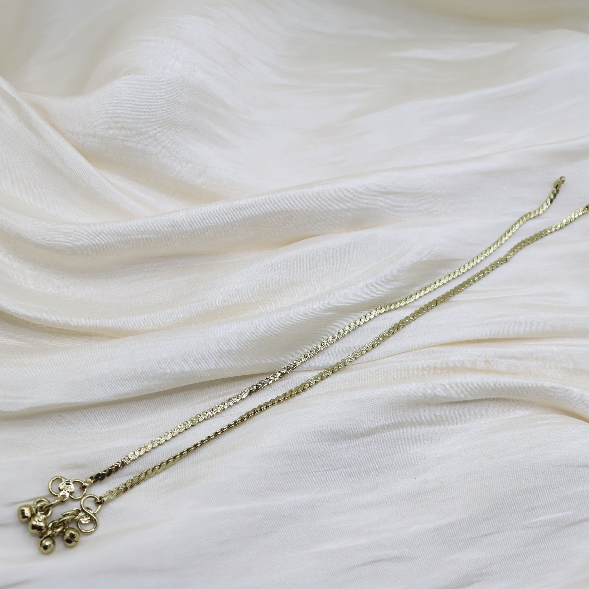 A delicate gold chain ankley with a small floral pendant, laid out diagonally on draped white fabric.