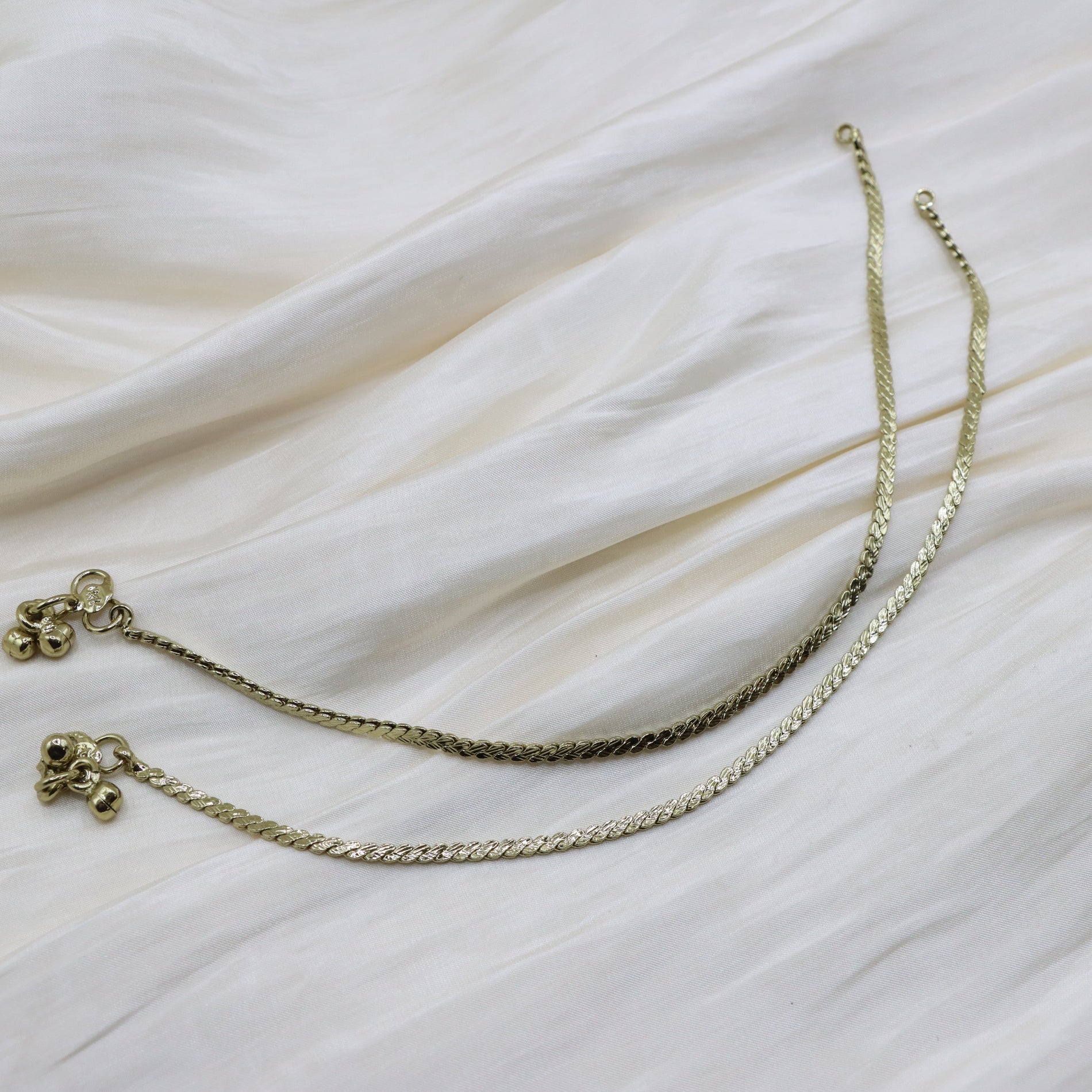 A gold-toned chain necklace with a textured woven design and small decorative charms, laid out on a draped white fabric background.