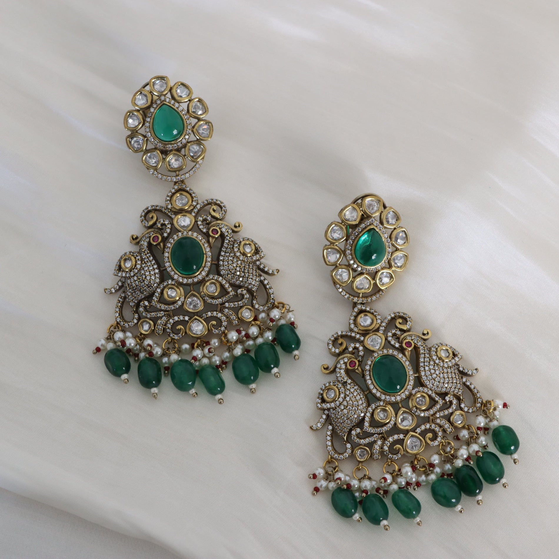 A pair of ornate gold-toned jhumka earrings with green stones and white faux pearls, displayed on a soft white fabric background.