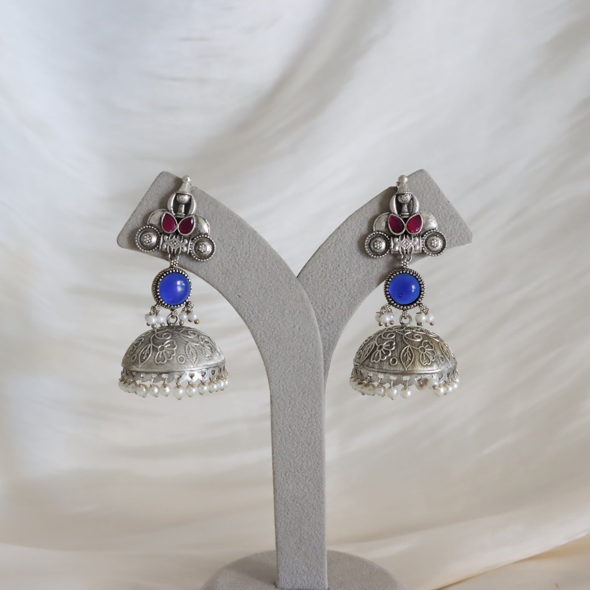 A pair of silver-toned Indian jhumka earrings with blue stone accents and small pearl bead borders, displayed on a gray jewelry stand against a soft white fabric background.