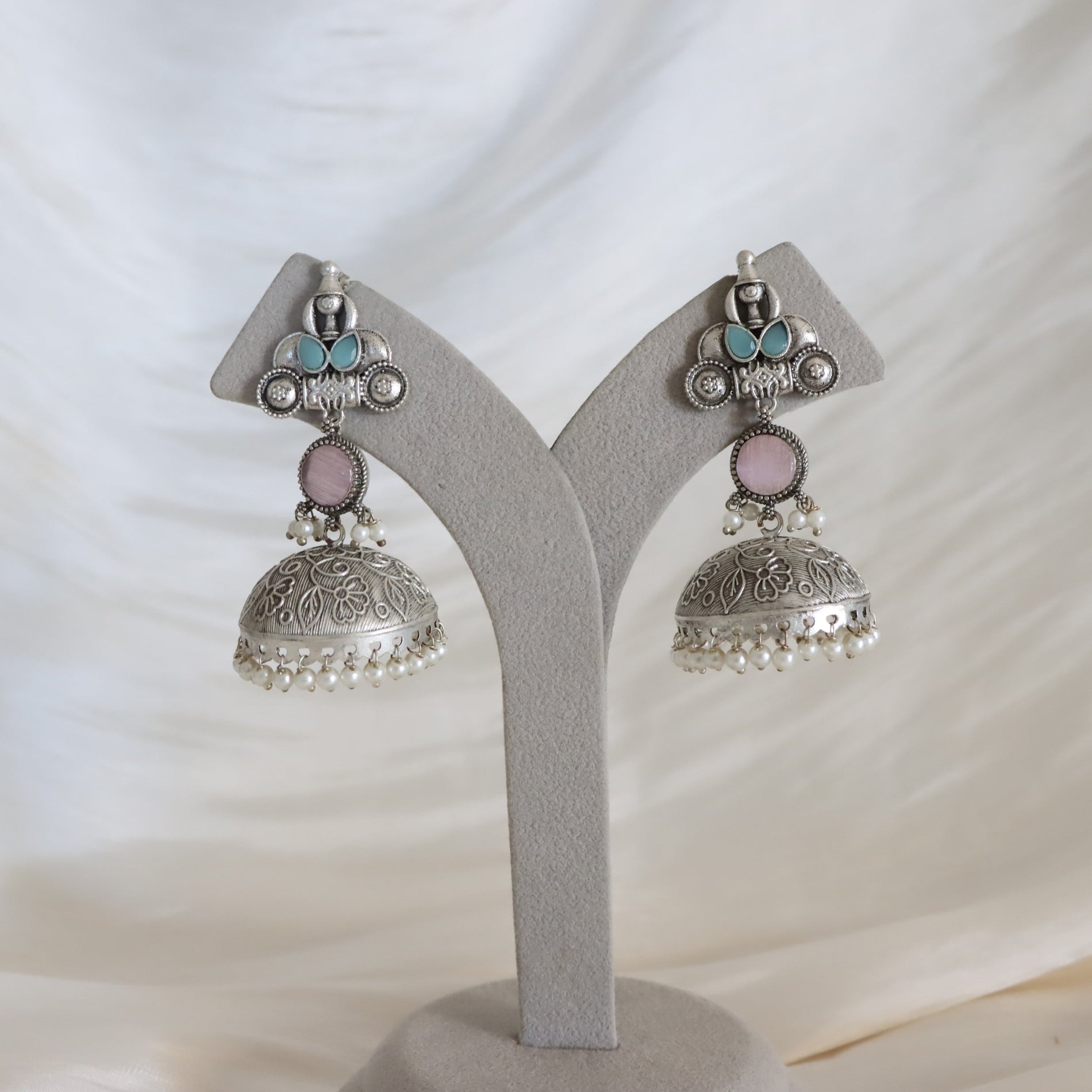 A pair of silver-toned Indian jhumka earrings with light blue and pink stones and white pearl bead fringe, displayed on a gray Y-shaped jewelry stand against a draped white fabric background.