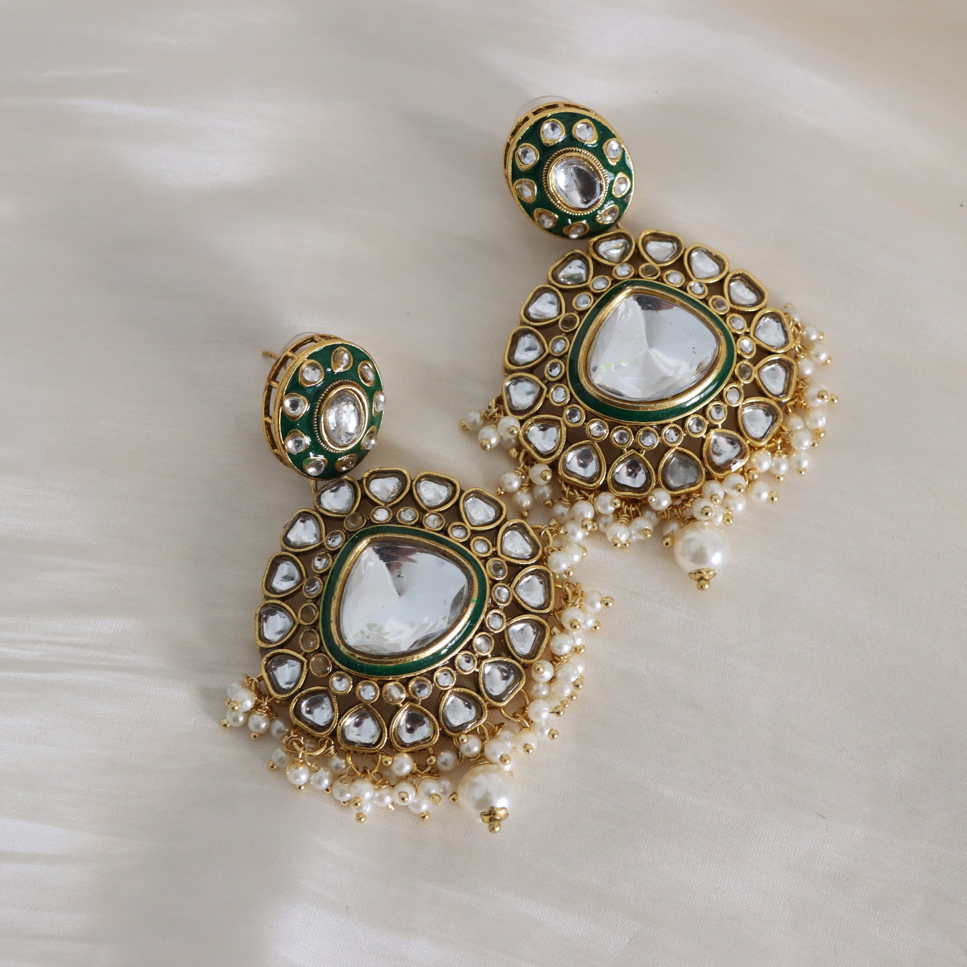 A pair of ornate gold-toned Indian Kundan earrings with green enamel accents, clear stone borders, and white faux pearl fringe, displayed on a soft white satin fabric background.