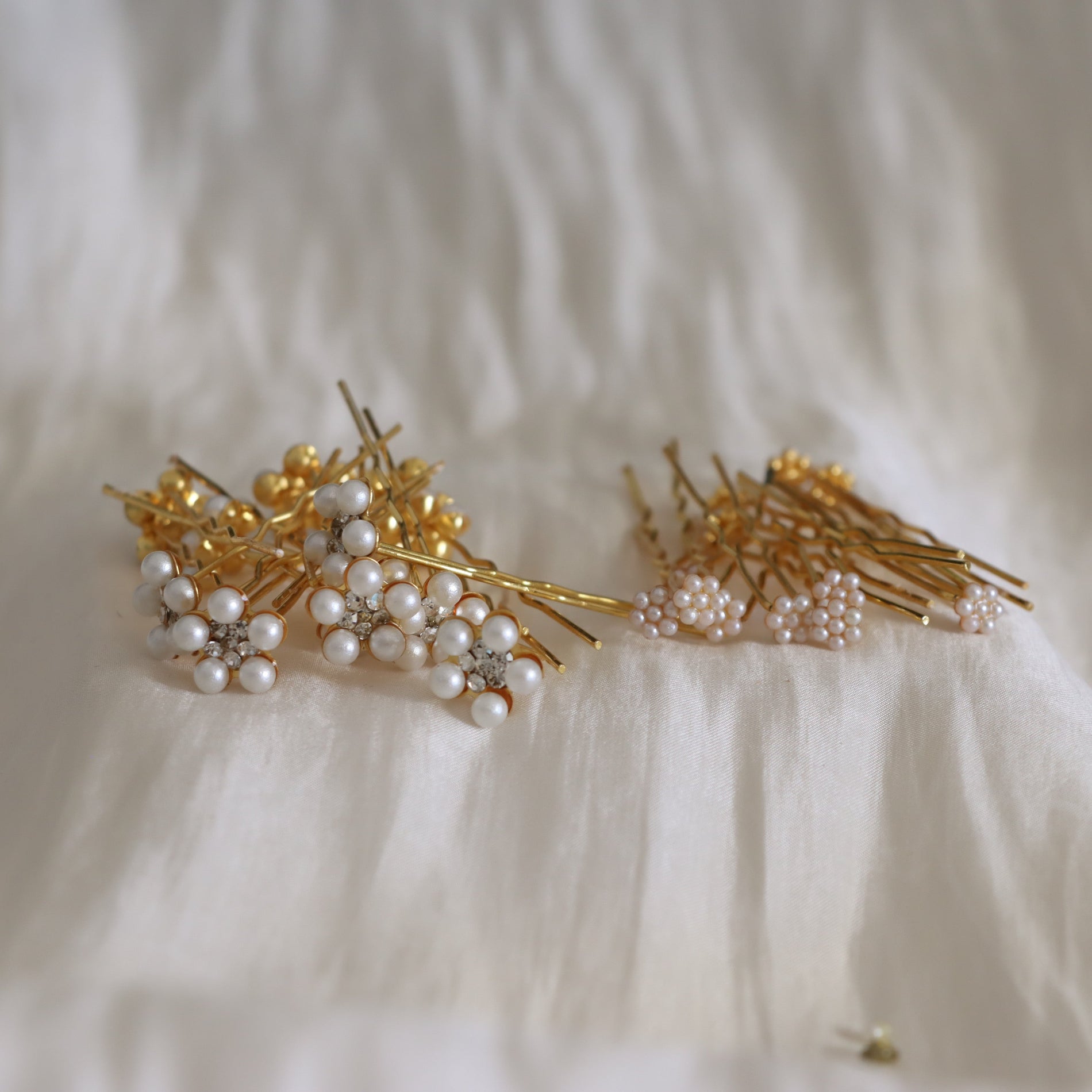 A set of gold hair pins decorated with clusters of white faux pearls and small clear rhinestones, arranged on a soft white textured fabric background.