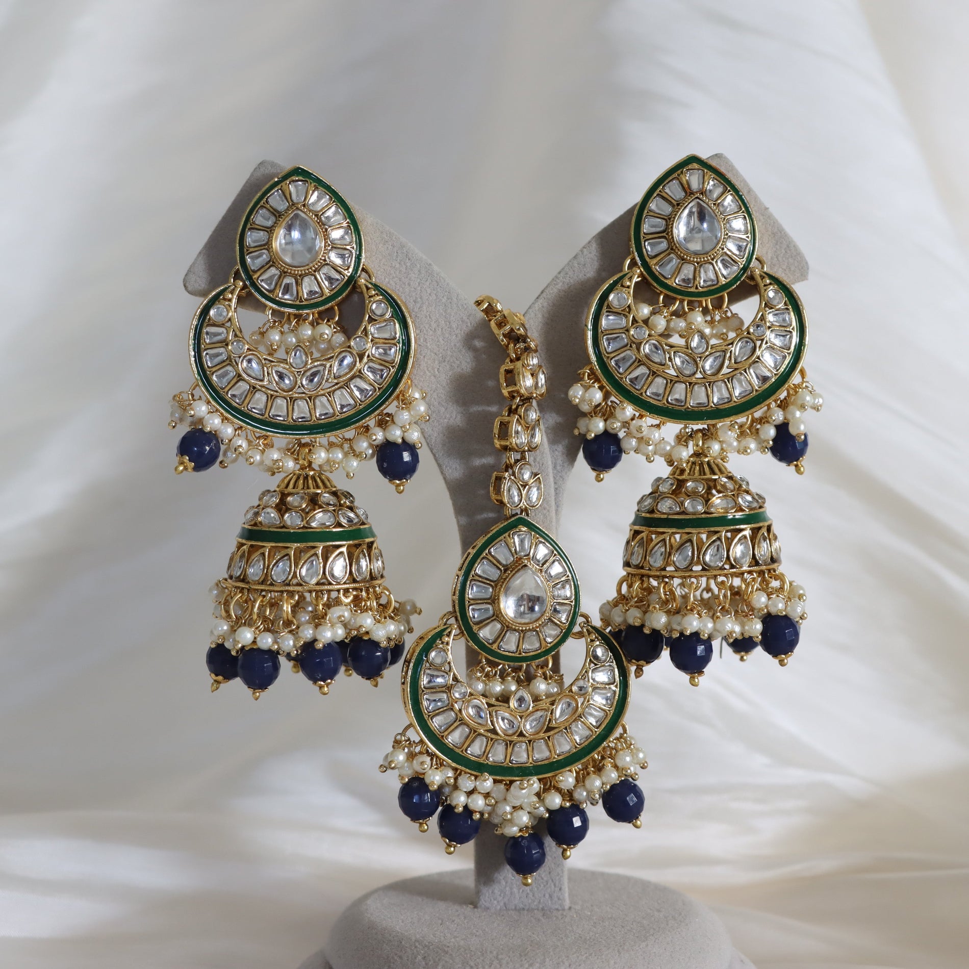 Gold-plated Indian jhumka earrings with green enamel, white faux pearls, and dark blue beads, displayed on a gray stand against a white draped fabric background.