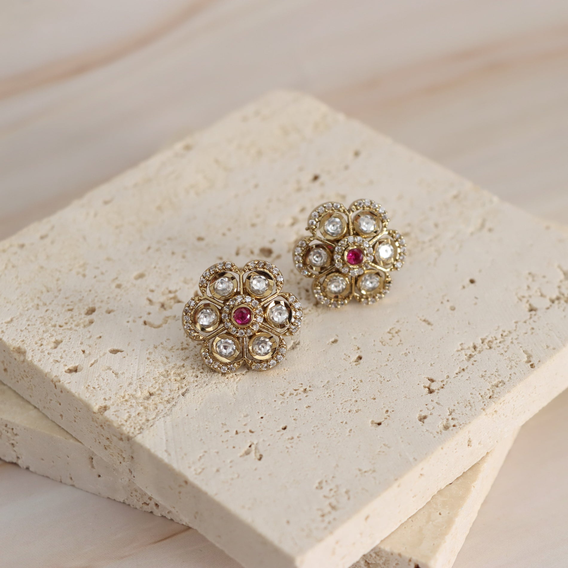 A pair of gold-toned floral stud earrings with clear rhinestones and a small red center stone, displayed on stacked beige stone tiles.