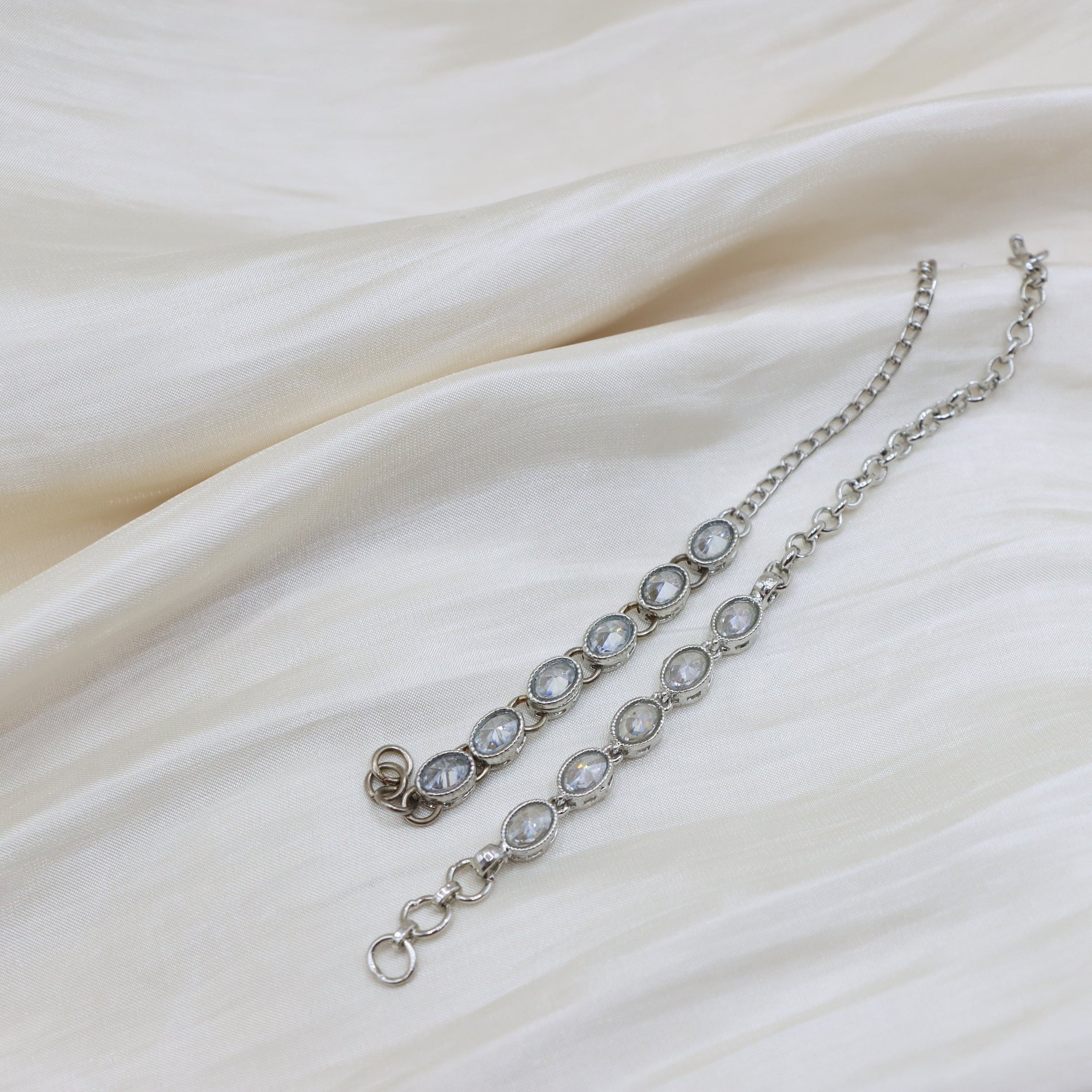 A silver-toned chain bracelet with oval clear gemstones, laid out on draped cream-colored fabric.