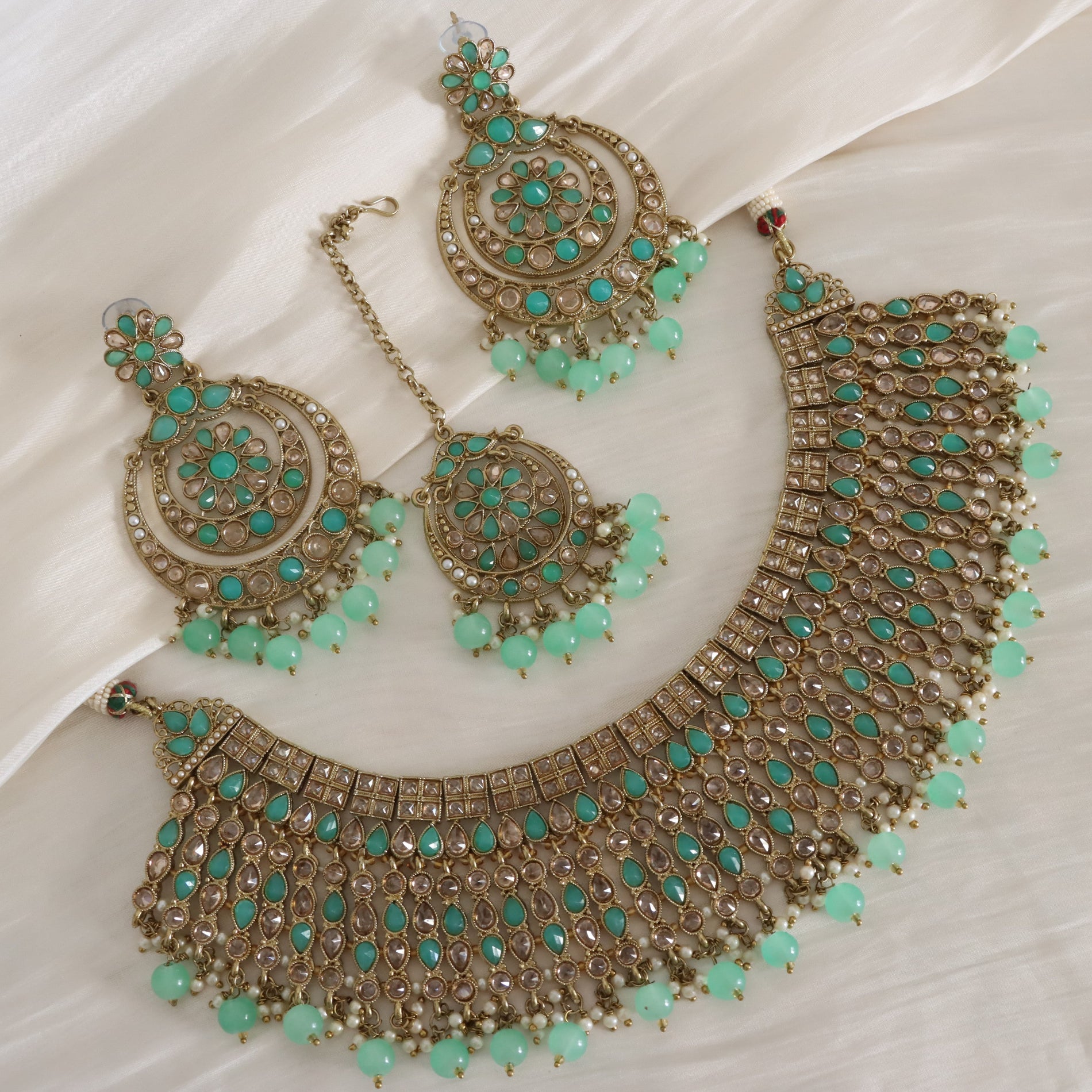 An ornate Indian ethnic jewelry set featuring a gold-plated necklace, matching earrings, and a maang tikka, embellished with turquoise green stones and bead drops, displayed on a white draped fabric background.