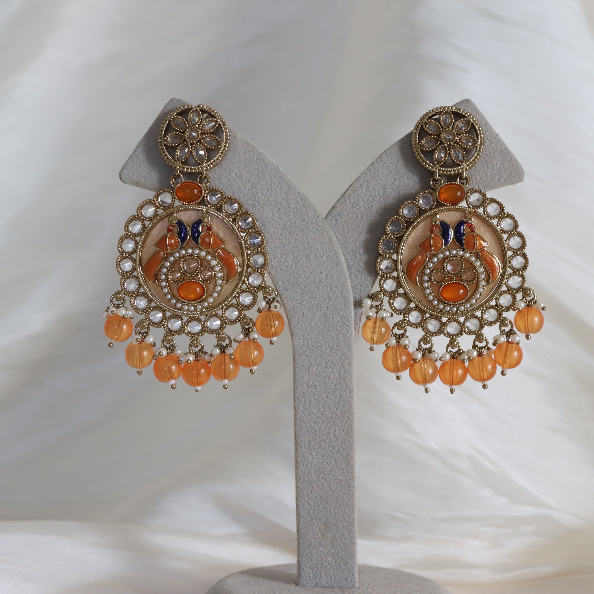 A pair of ornate gold-toned Indian jhumka earrings with orange glass beads, clear rhinestones, and blue butterfly details, displayed on a gray jewelry stand against a white fabric background.