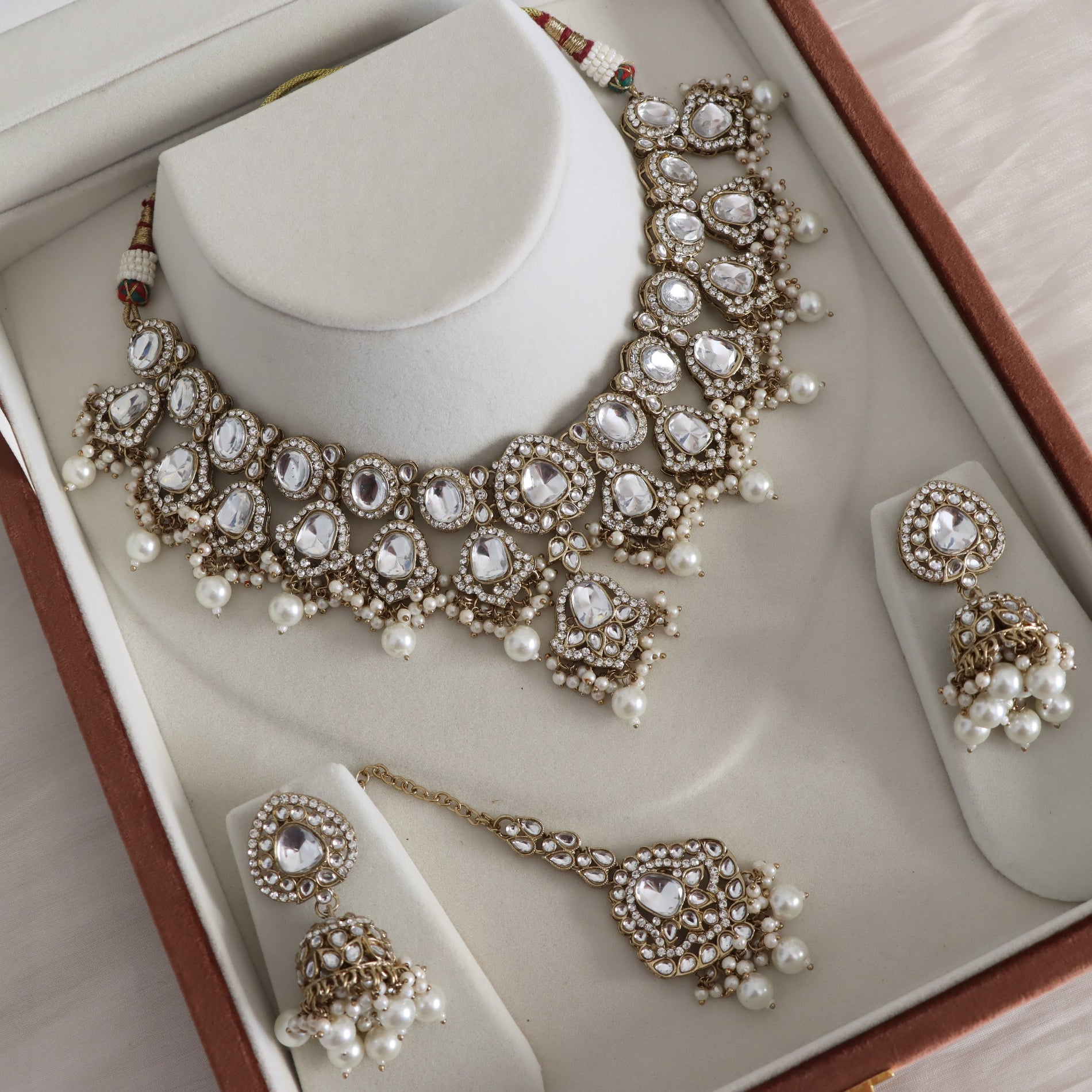 An ornate Indian ethnic jewelry set displayed inside its brown presentation box. The set includes a choker necklace, a matching pair of dangle earrings, and a maang tikka, all decorated with gold-toned metalwork, clear stones, and white faux pearls.