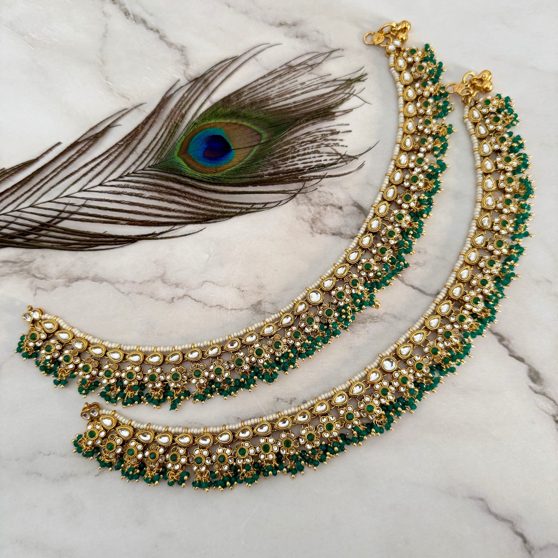 Ornate gold and green anklet on a marble surface with a peacock feather.