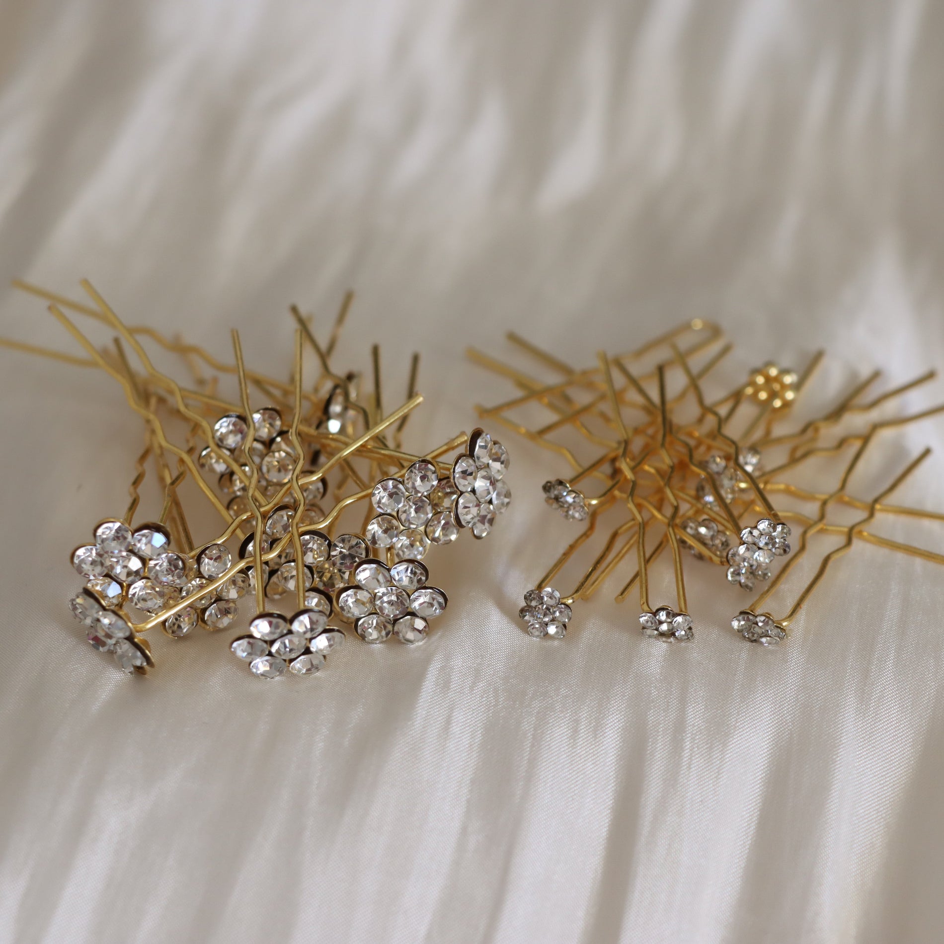Two gold hair pins adorned with clear rhinestones, arranged on a white satin fabric background.