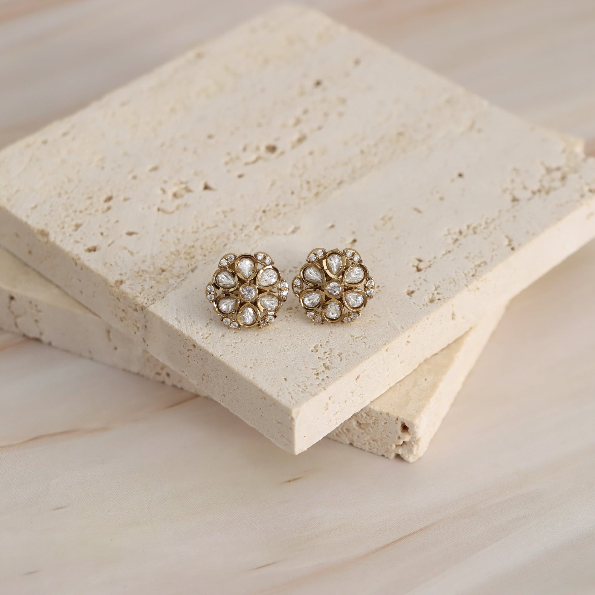 A pair of gold-toned stud earrings with clear teardrop-shaped crystals arranged on a textured beige stone tile over a light wood surface.