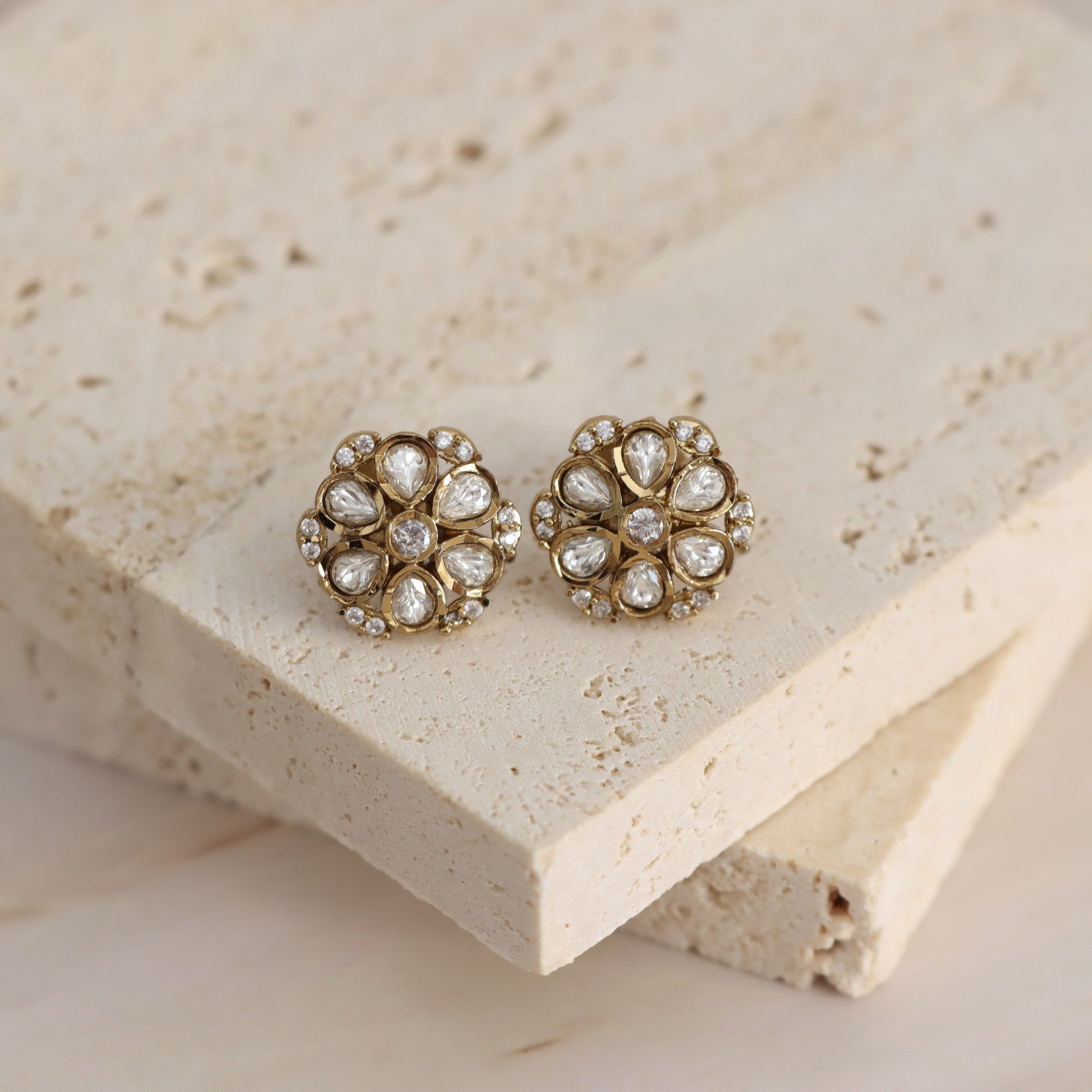 A pair of gold-toned floral stud earrings with clear teardrop-shaped stones and small pearl-like accents, displayed on stacked beige stone blocks.