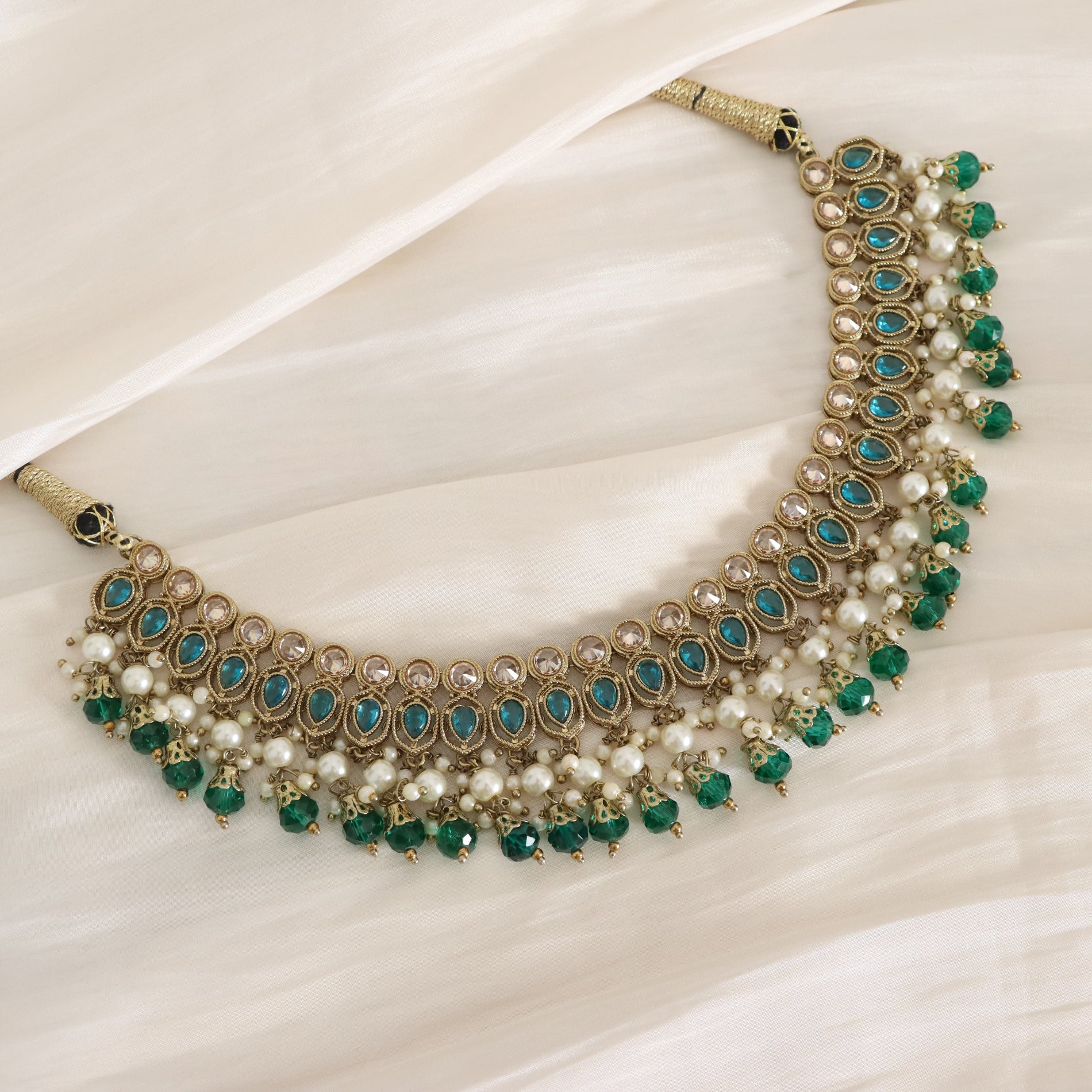 An ornate gold-toned statement necklace featuring rows of white faux pearls, green faceted beads, and turquoise-blue teardrop stones, displayed on draped cream fabric.