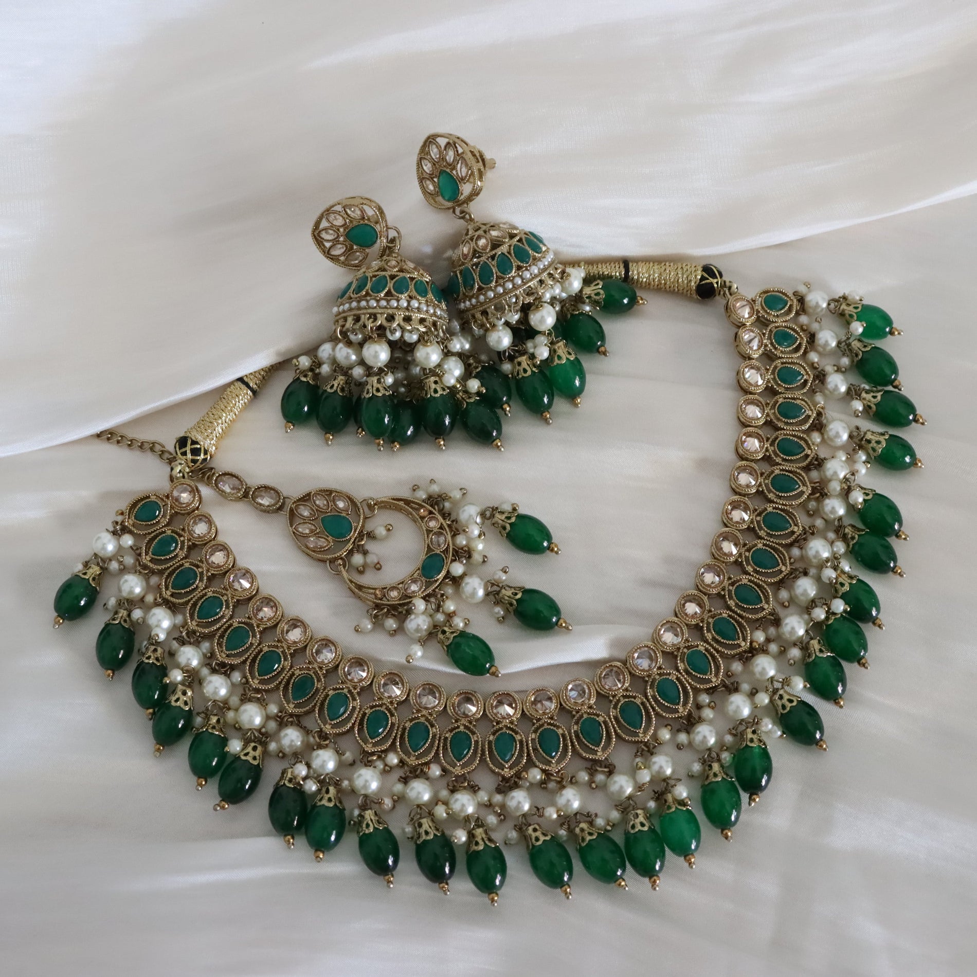 An ornate traditional Indian jewelry set featuring a statement necklace, matching earrings, and a curved bracelet, decorated with green stones, white faux pearls, and gold-plated filigree work, displayed on a white satin fabric background.