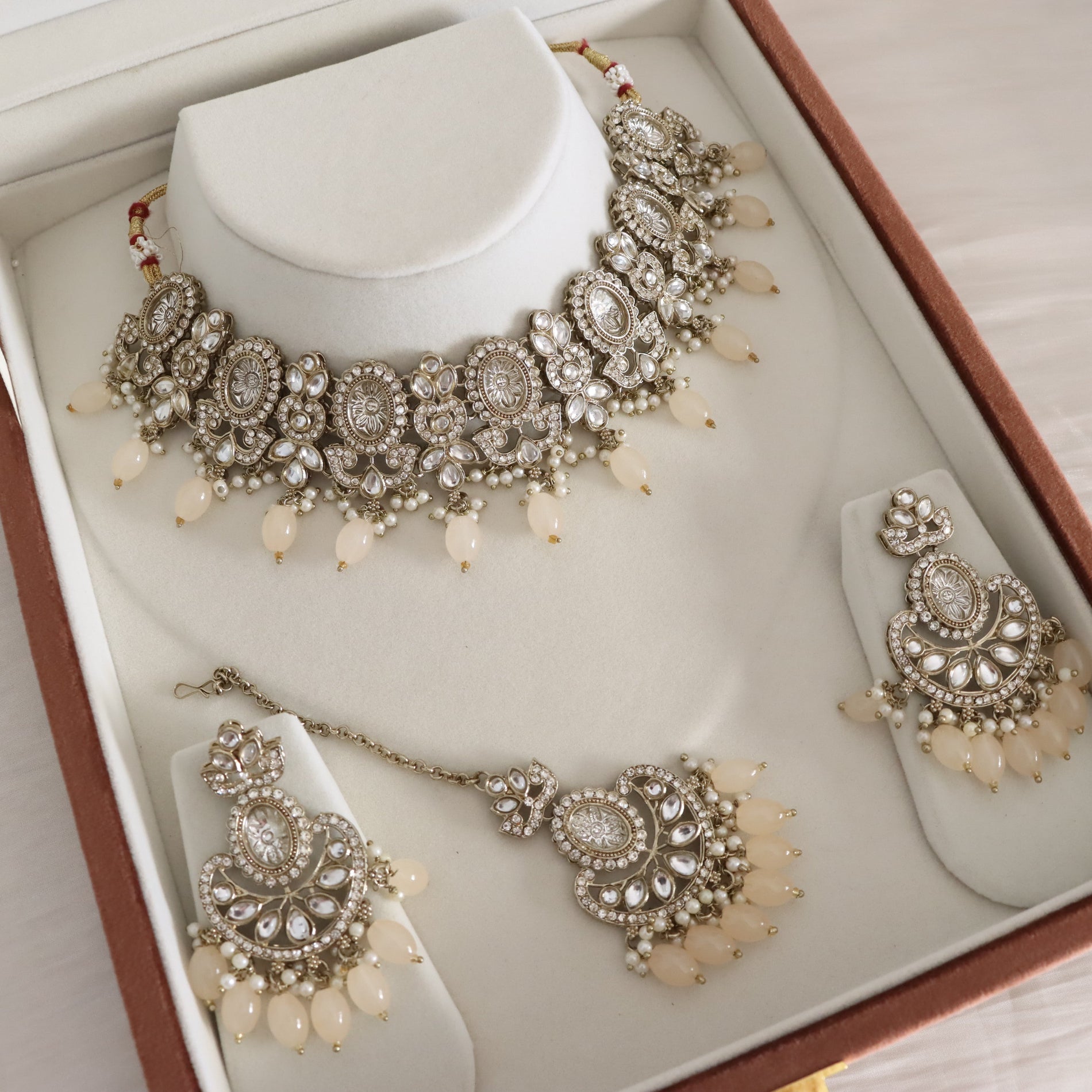 A traditional Indian bridal jewelry set displayed inside its brown presentation box. The set includes a silver-tone Kani necklace with cream pearl beads, matching Kara earrings, and a Kahan choker bracelet, all embellished with clear stones.