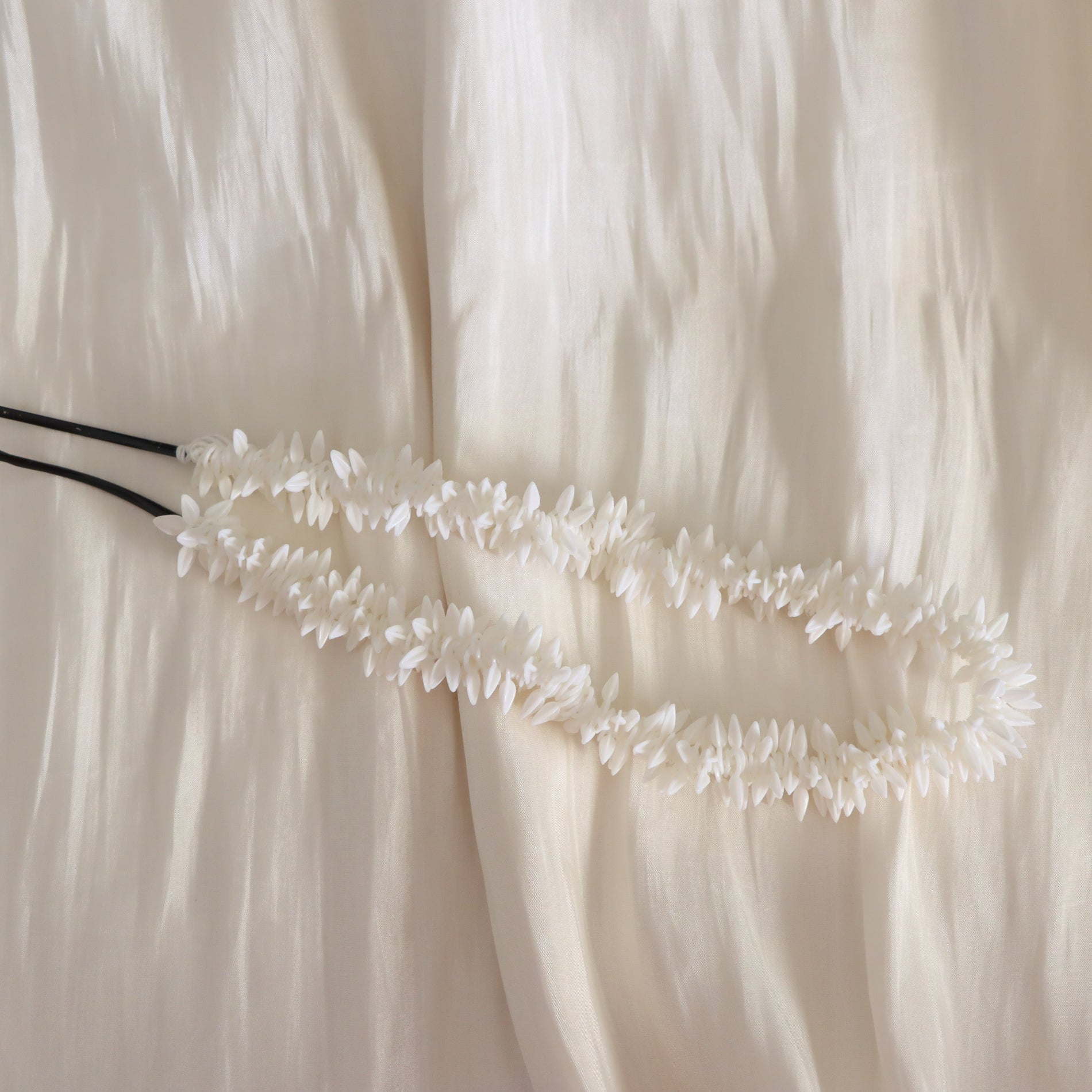 A white floral lei necklace with black elastic cords, laid flat on a textured, cream-colored fabric background.