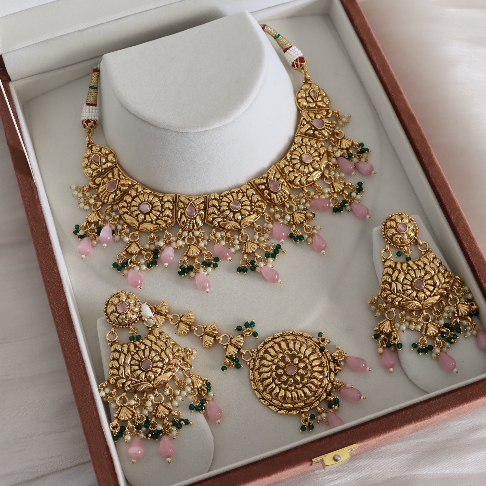 Gold-toned traditional Indian jewelry set with a necklace, earrings, and a round pendant, decorated with pink stones and green bead accents, displayed on a white jewelry stand inside an open presentation box.
