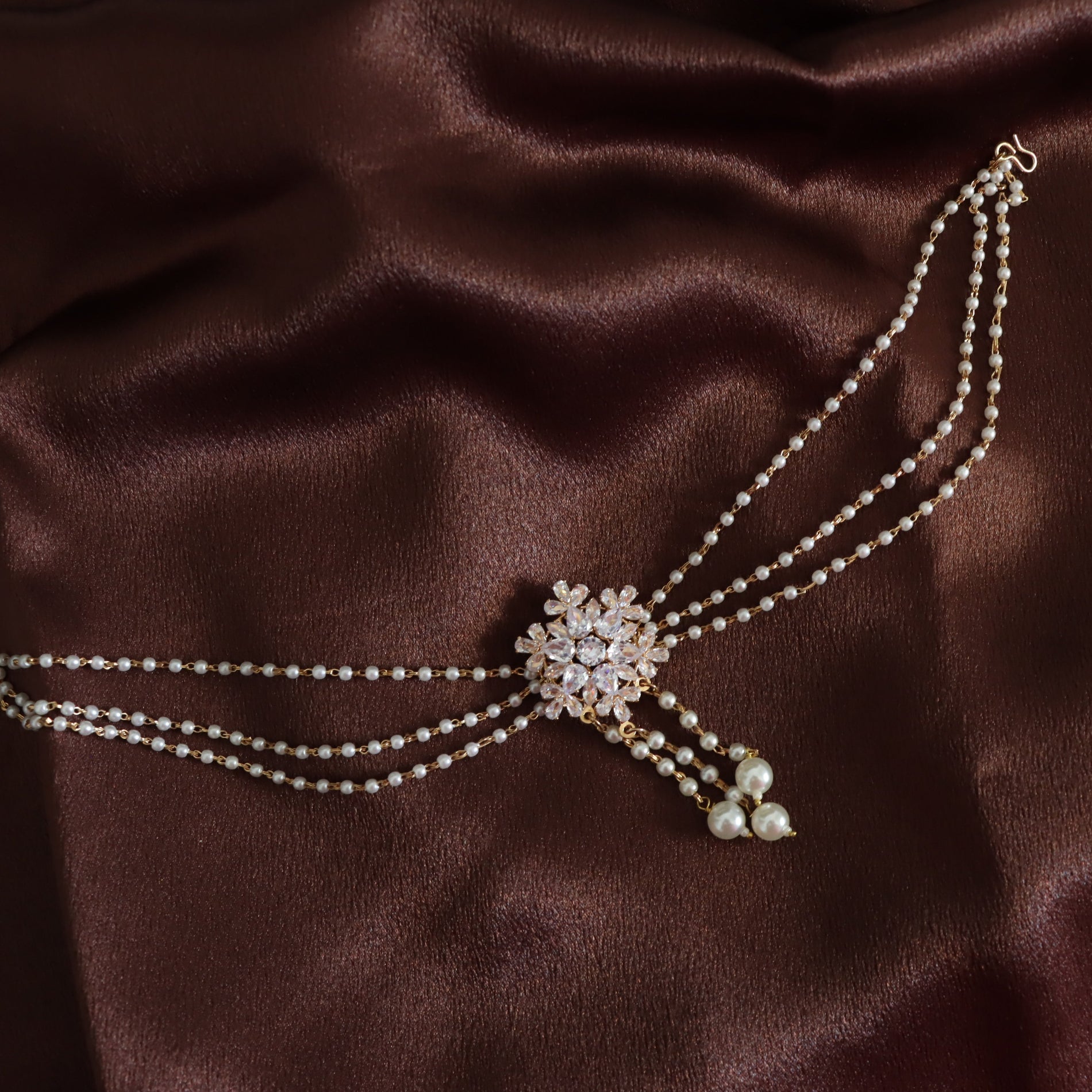 A double-strand pearl choker necklace with a central crystal-embellished floral brooch and dangling pearl accents, displayed on a draped dark brown satin fabric.