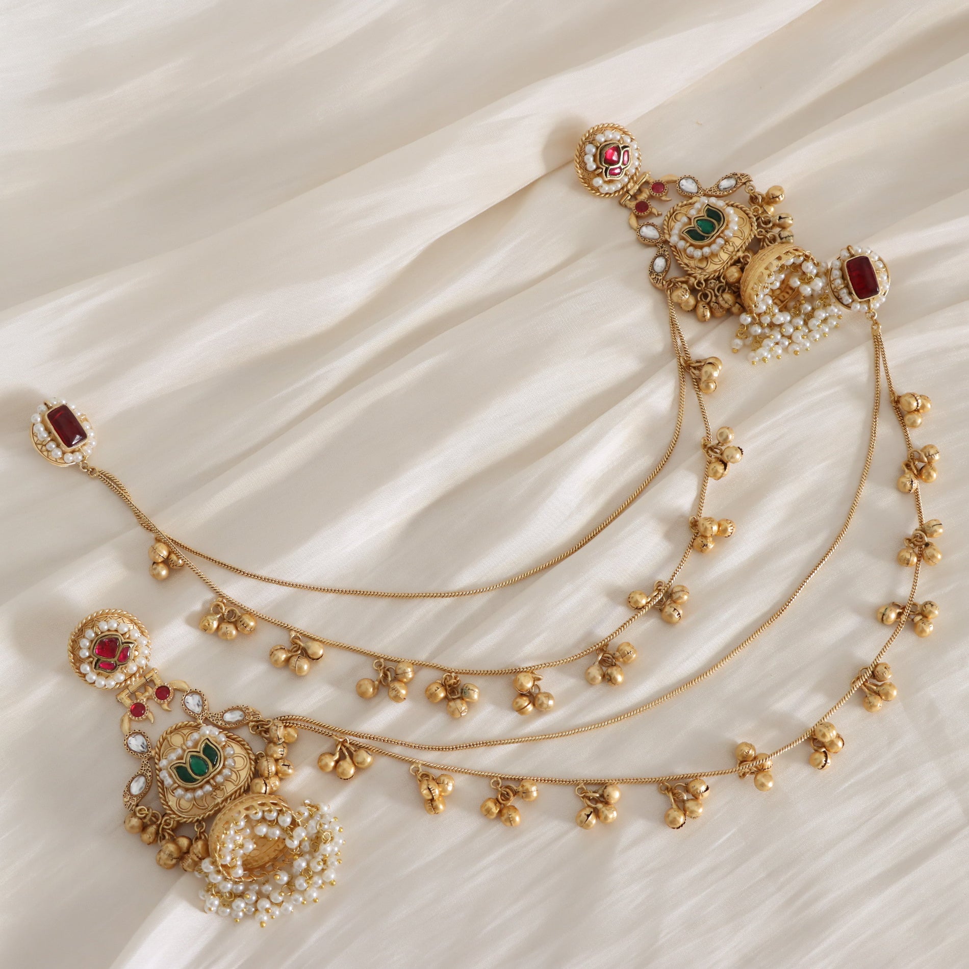 An ornate gold-toned traditional jewelry set with intricate floral motifs, white stones, and red and green gemstone accents, arranged on a draped cream fabric background.