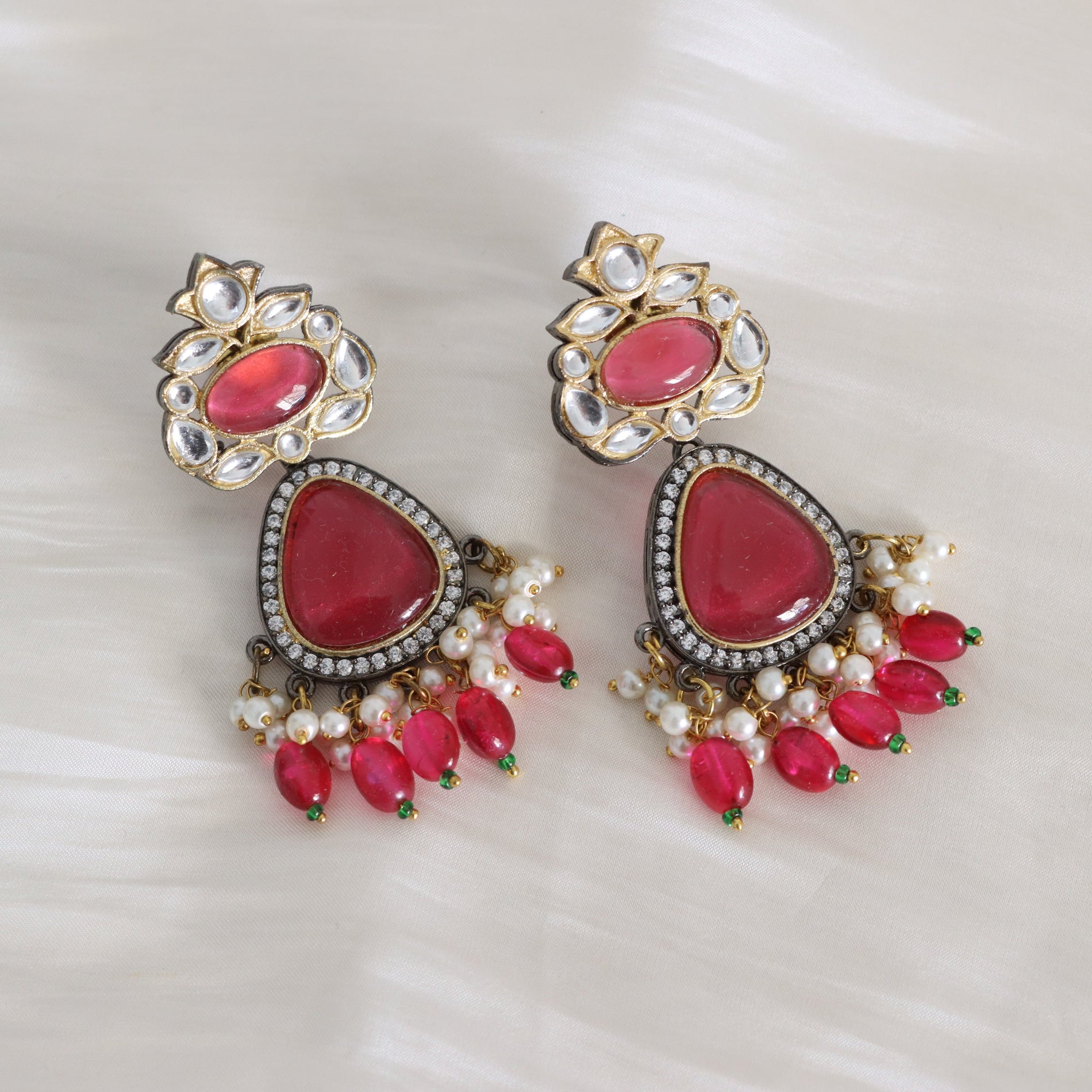 A pair of gold-toned Indian jhumka earrings with large pink teardrop-shaped stones, white faux pearl bead chains, and small green accent beads, displayed on a soft white fabric background.