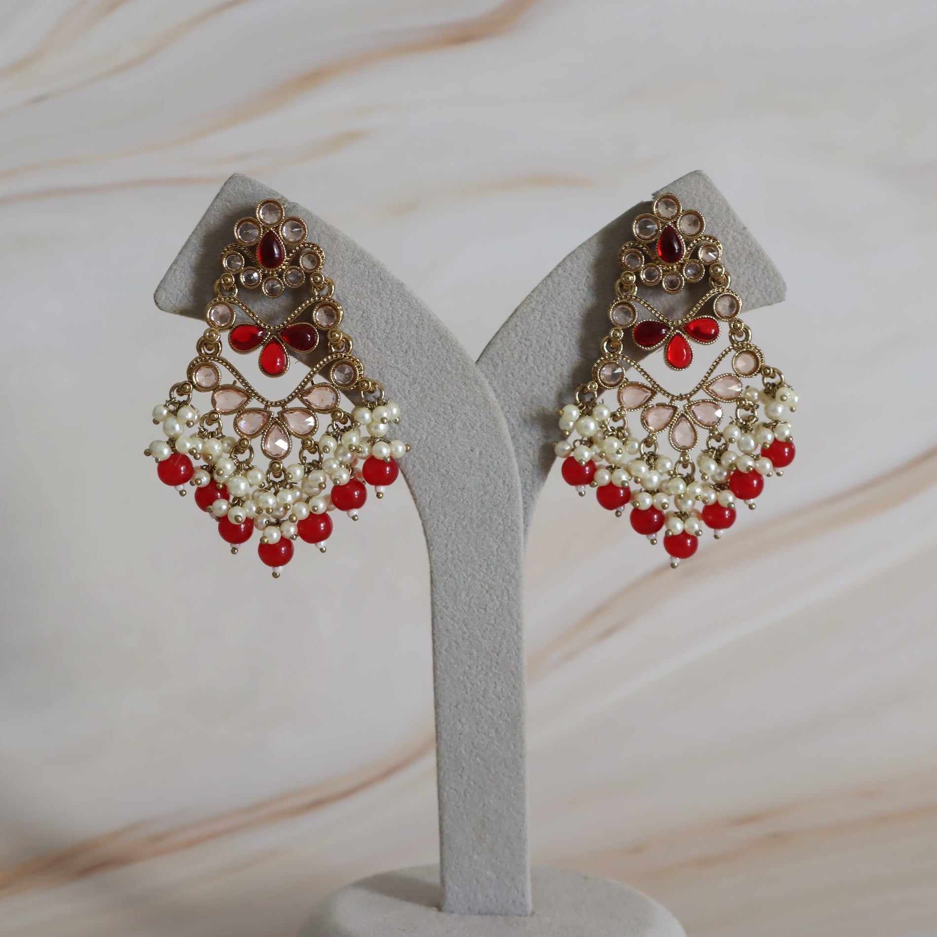Pair of ornate earrings with red and pearl-like stones on a gray stand against a light background