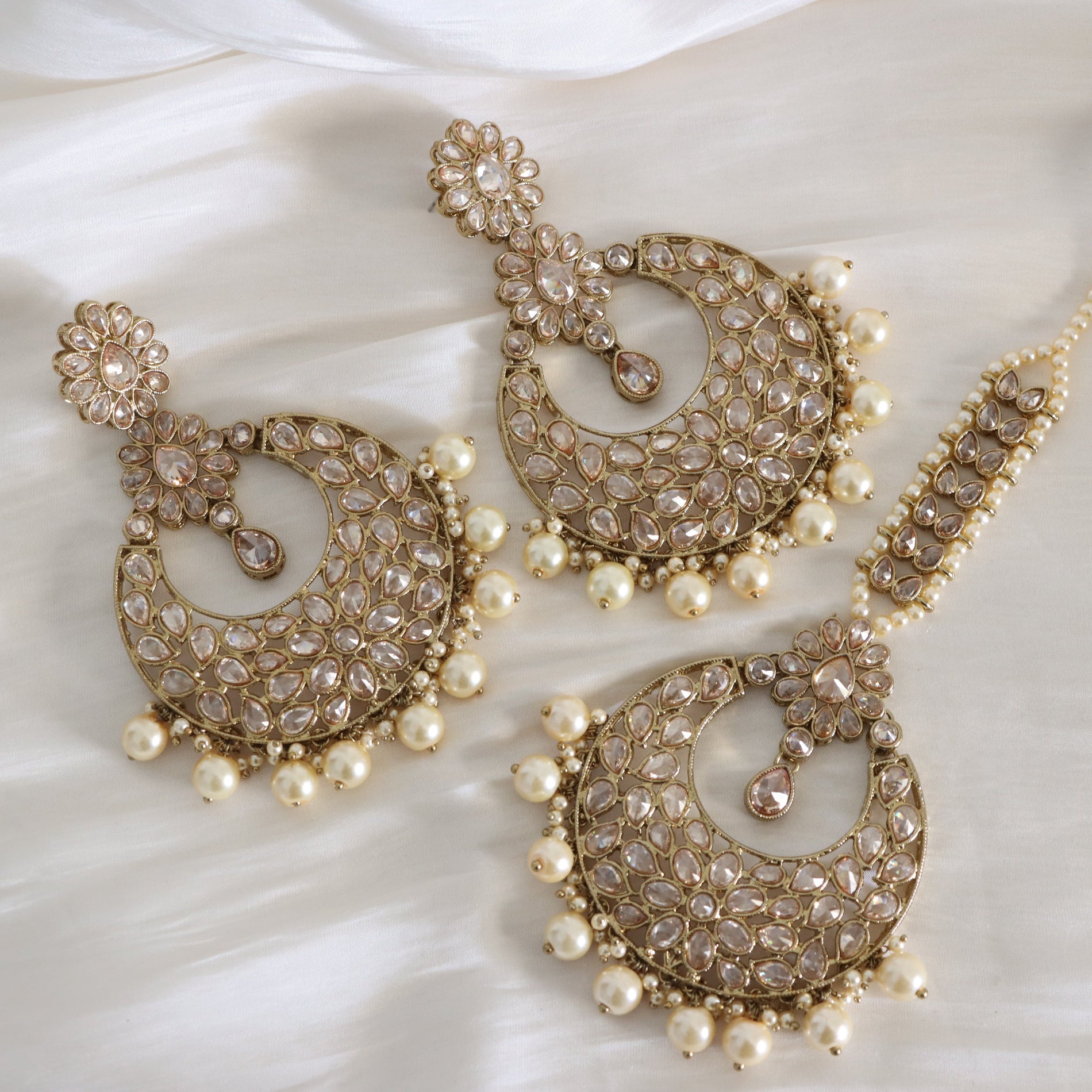 A set of ornate Indian jhumka earrings and a matching maang tikka laid out on a white satin fabric background. The jewelry is made of gold-plated metal heavily embellished with clear crystals and decorated with rows of white faux pearls.