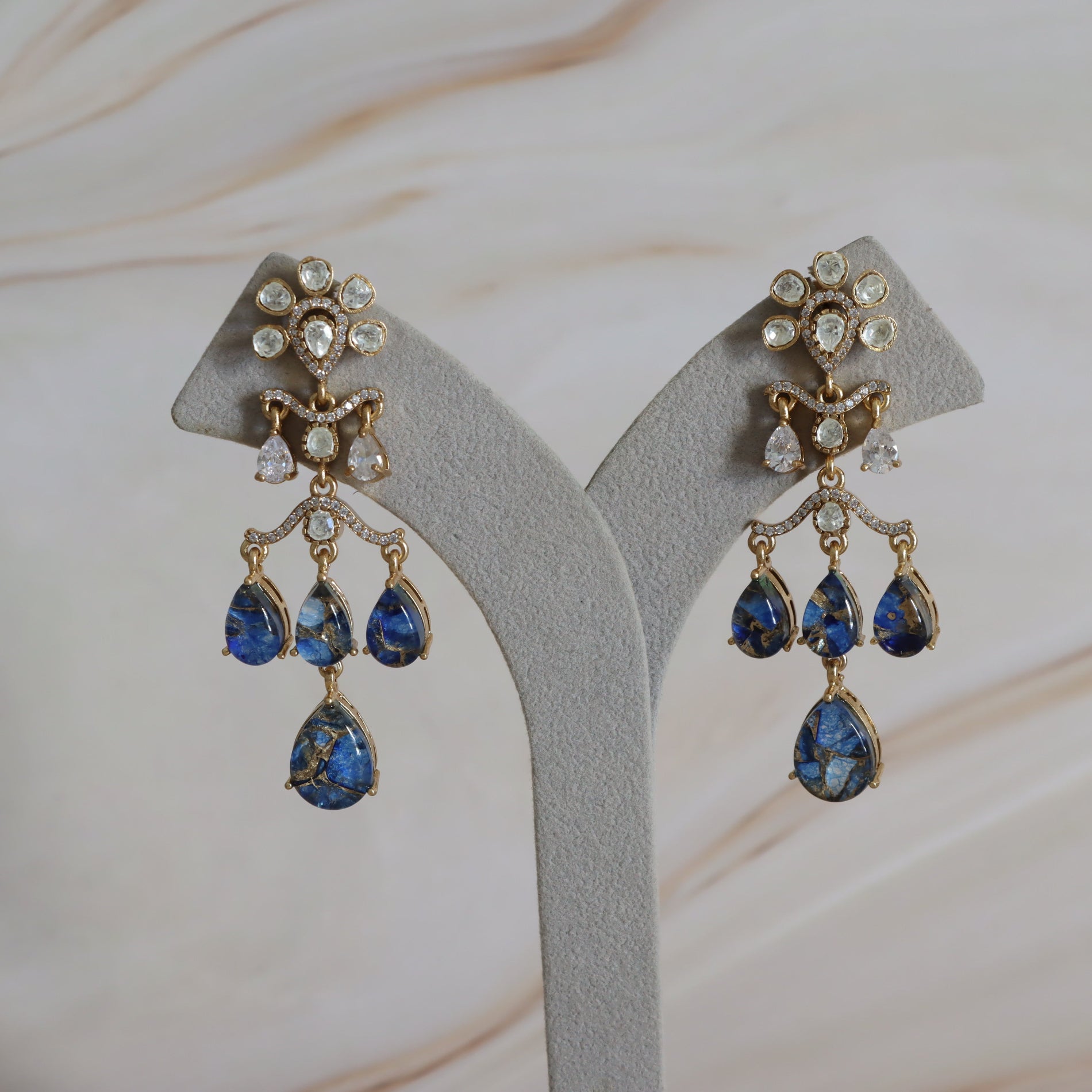 Pair of gold earrings with blue teardrop stones on a gray stand.