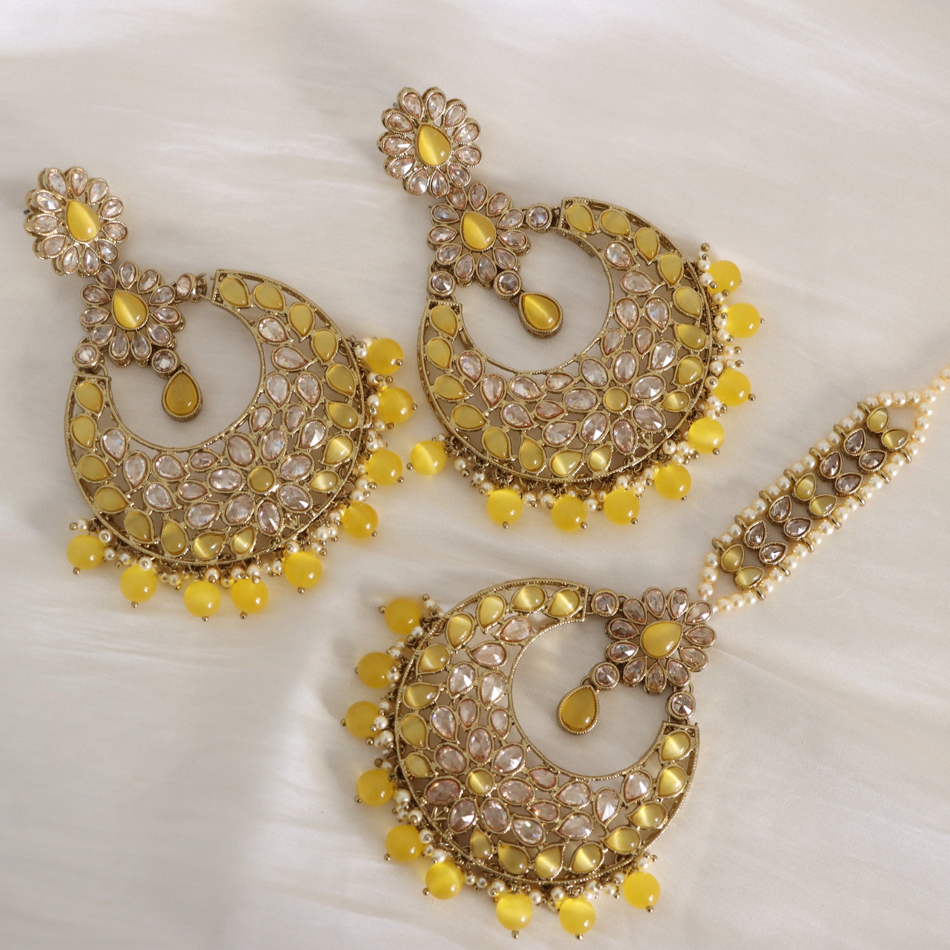 A traditional Indian gold-toned jewelry set featuring a crescent-shaped jhumka earring, a matching maang tikka headpiece, and a long pearl-beaded chain, all embellished with yellow stones and clear crystals on a white fabric background.