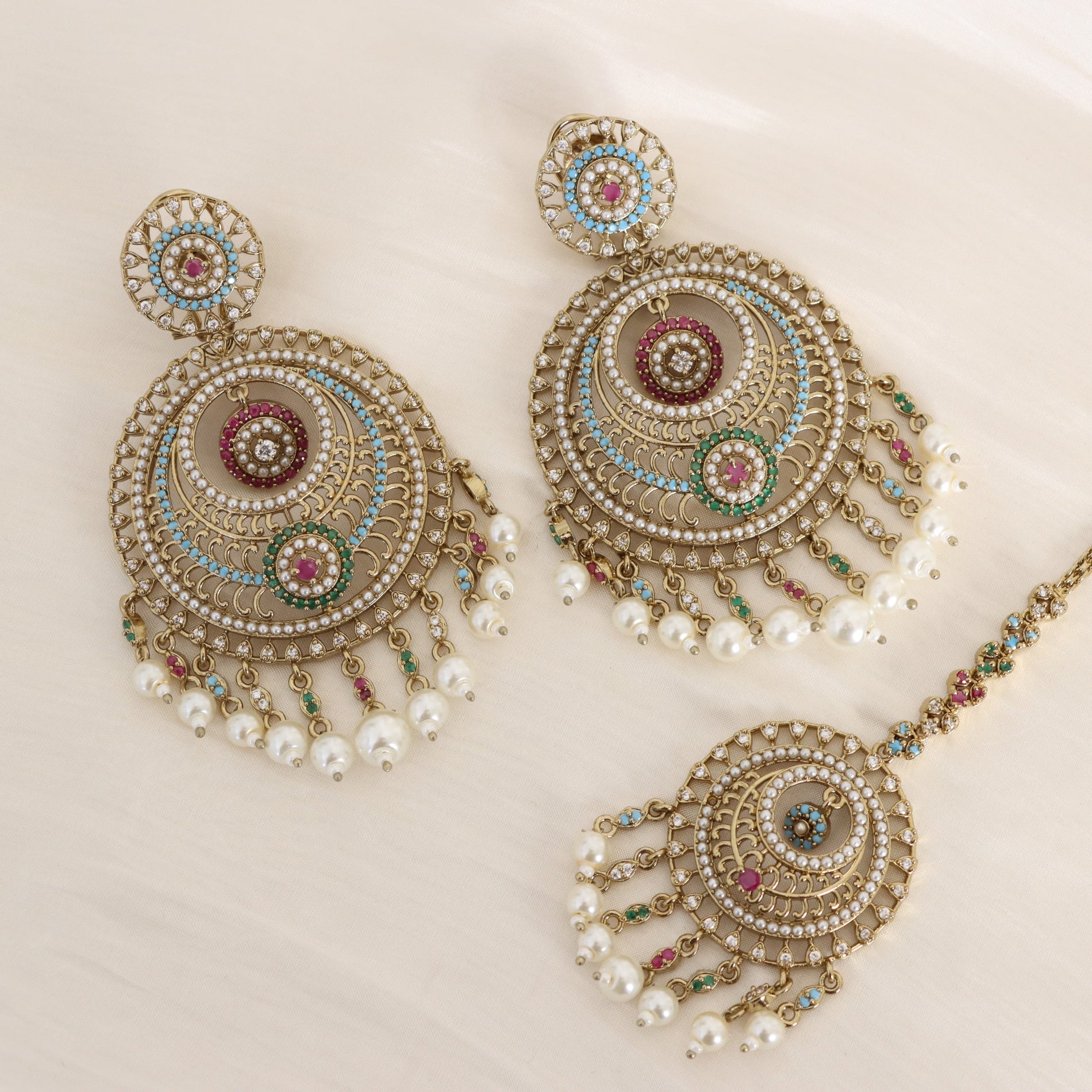 Gold-toned Indian jhumka earrings with matching necklace and a nose chain, decorated with white faux pearls, turquoise blue, pink, and green stones on a light cream background.