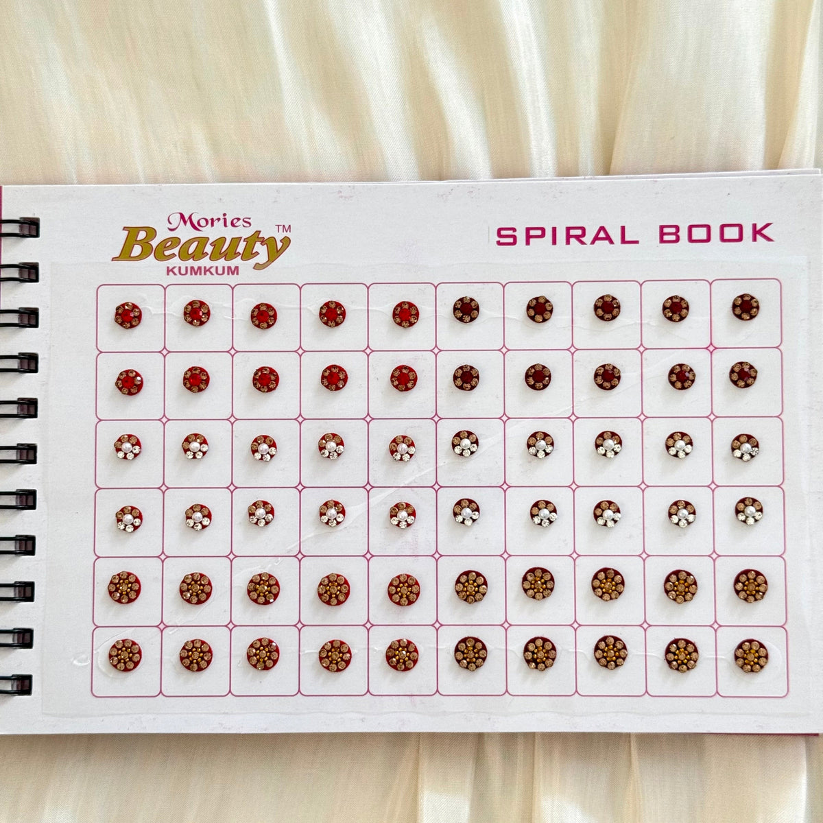 A spiral-bound beauty rhinestone display book showing rows of small red and gold faceted gems on a white page, with the text 'Mories Beauty KUMKUM SPIRAL BOOK' visible.