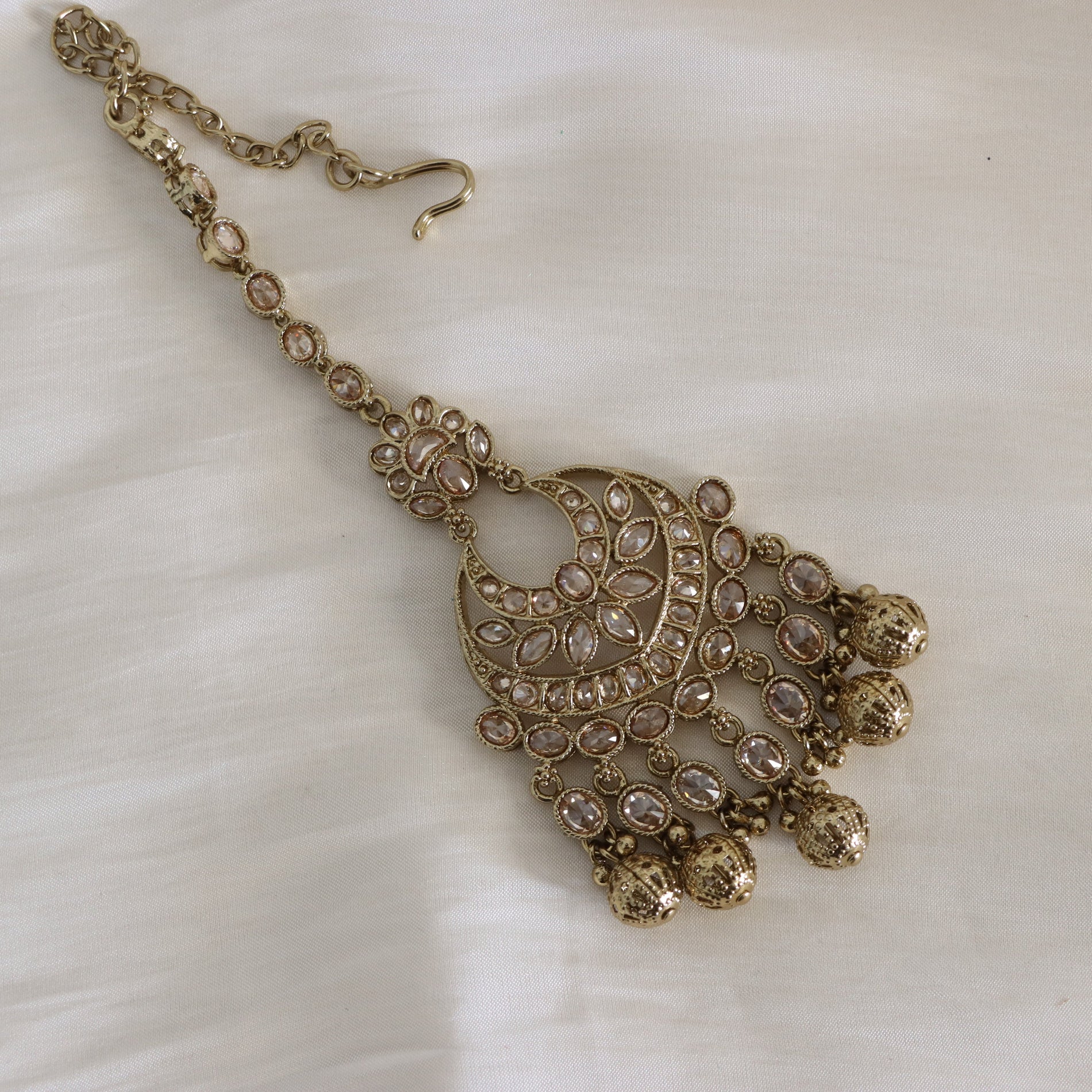 An ornate gold-toned traditional Indian necklace with a large crescent-shaped pendant decorated with clear stones and hanging bell-shaped charms, displayed on a white fabric background.