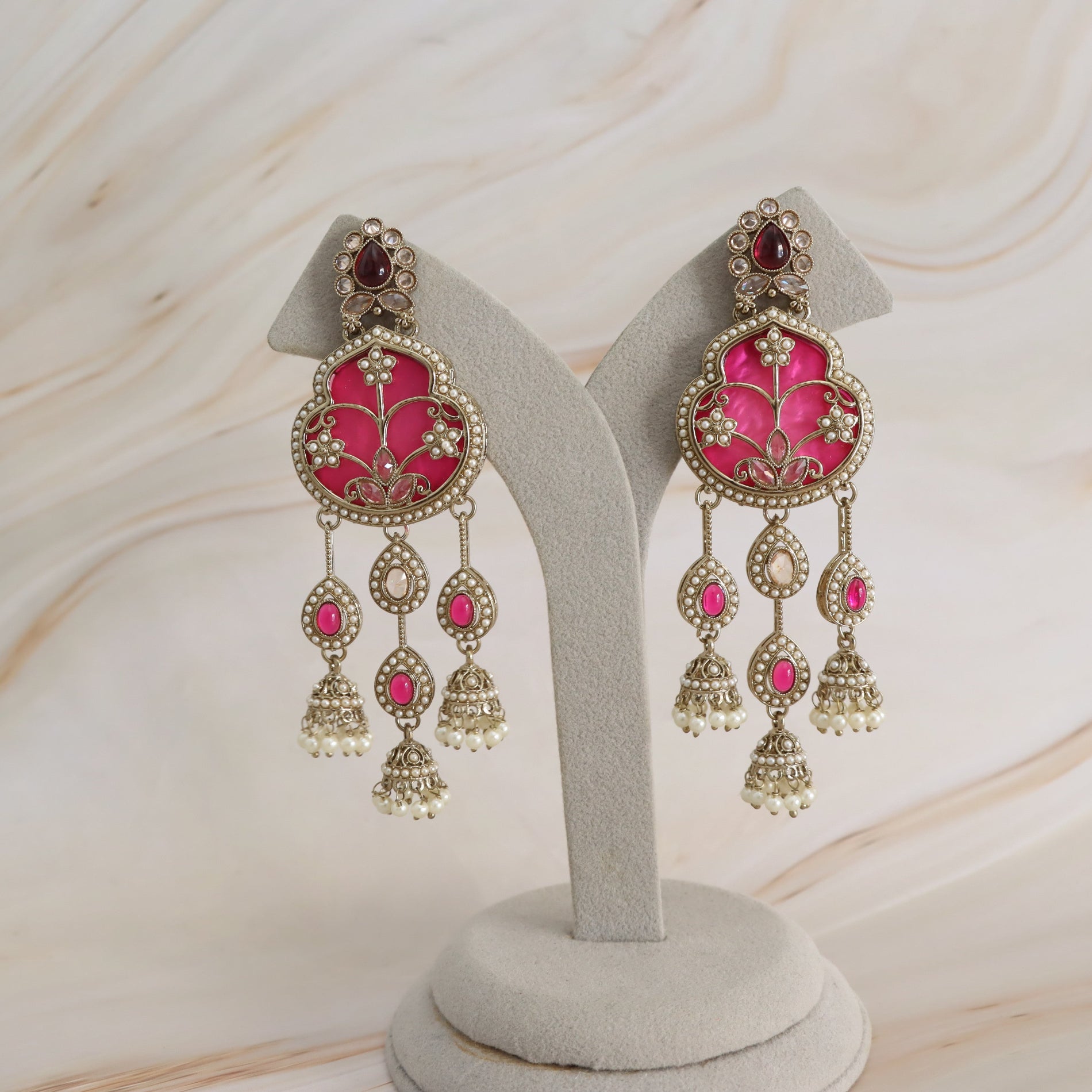 A pair of ornate gold-toned dangle earrings with bright pink enamel floral panels and small pearl-like bead details, displayed on a light gray jewelry stand against a soft beige marble-patterned background.
