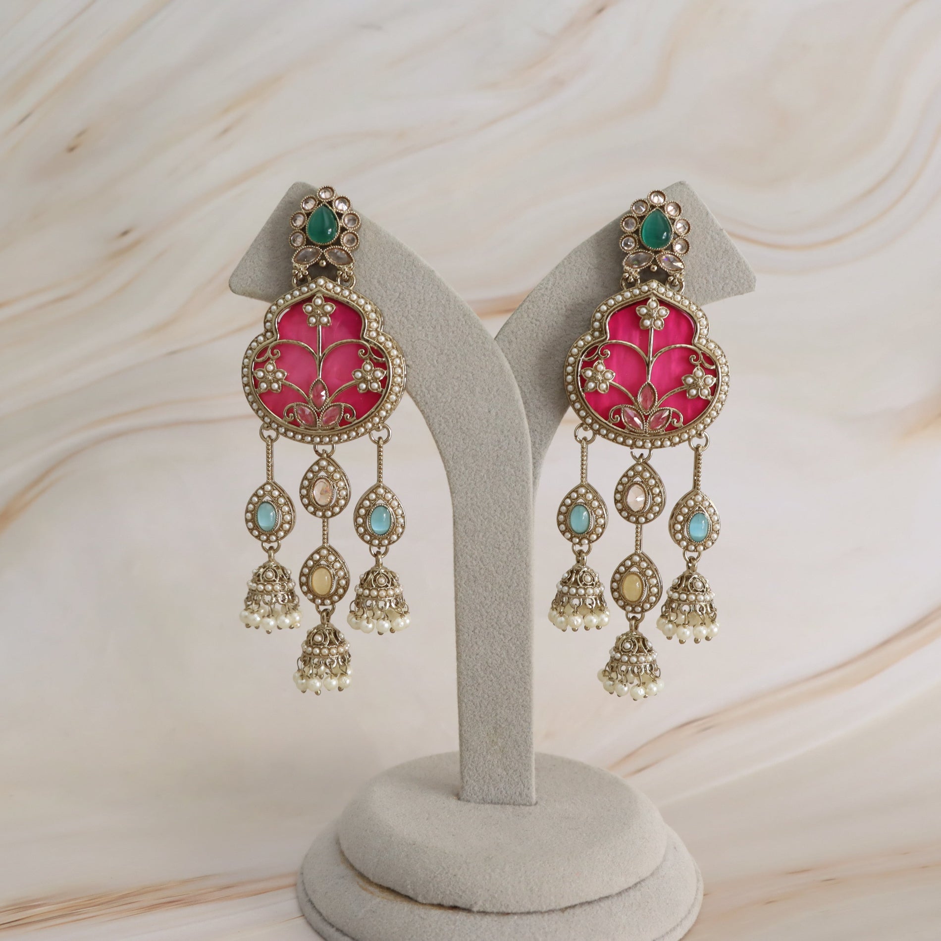 A pair of ornate gold-toned dangle earrings with bright pink floral inlay, green stones at the top, and small turquoise and gold accents, displayed on a light gray jewelry stand against a soft beige marble background.