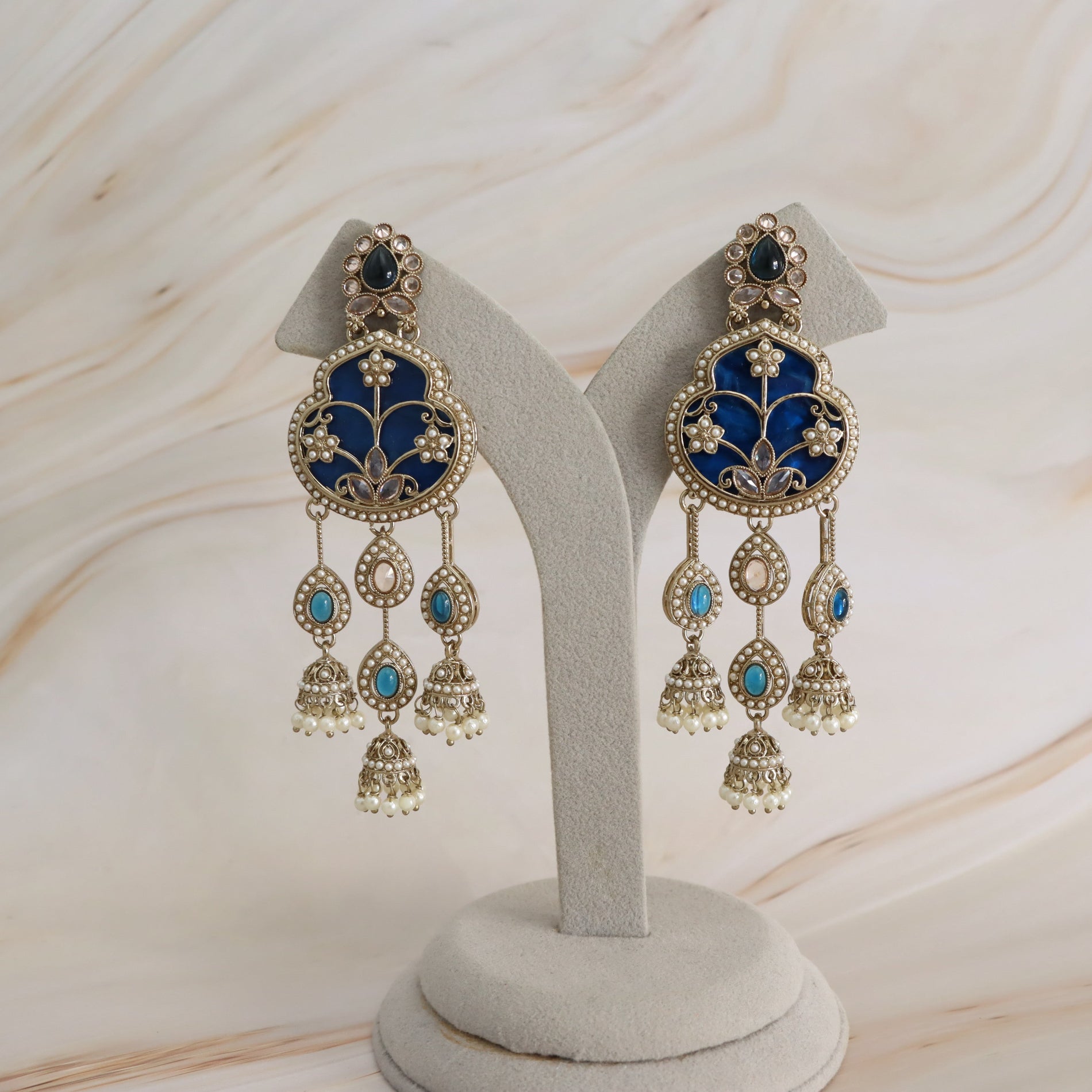 A pair of ornate gold-toned Indian jhumka earrings with deep blue enamel panels and small turquoise accents, displayed on a light gray jewelry stand against a cream marble background.