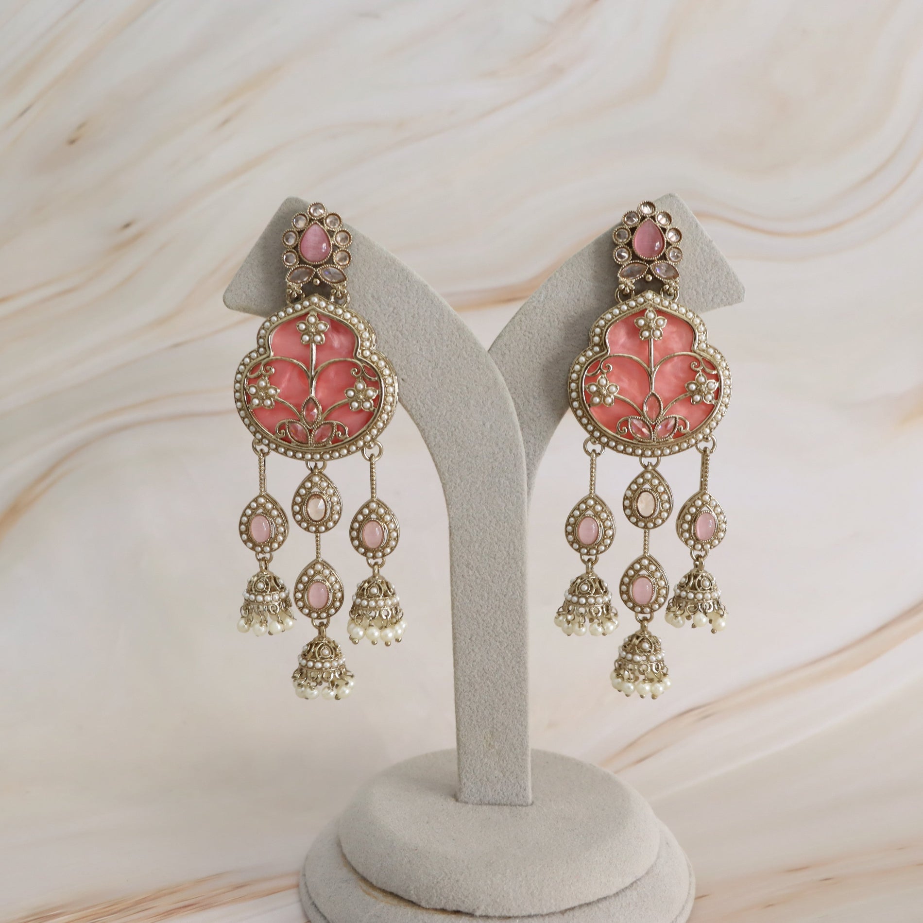 A pair of ornate pink and gold Indian jhumka earrings displayed on a light gray velvet jewelry stand against a light beige marble background.