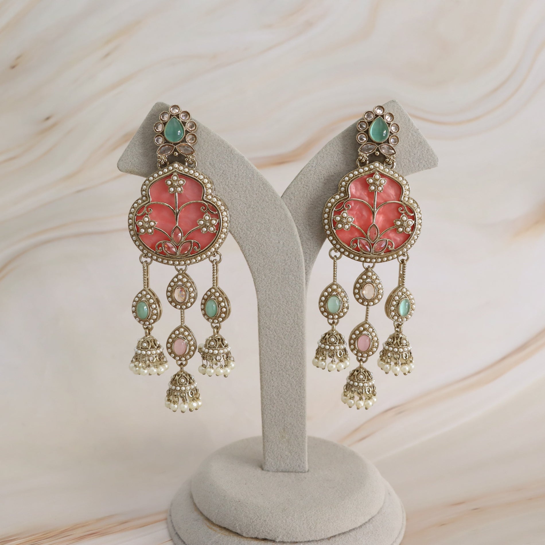 A pair of ornate gold-toned dangle earrings with coral-pink floral enamel, green stones, and dangling bell-shaped charms, displayed on a light gray jewelry stand against a soft beige marble background.