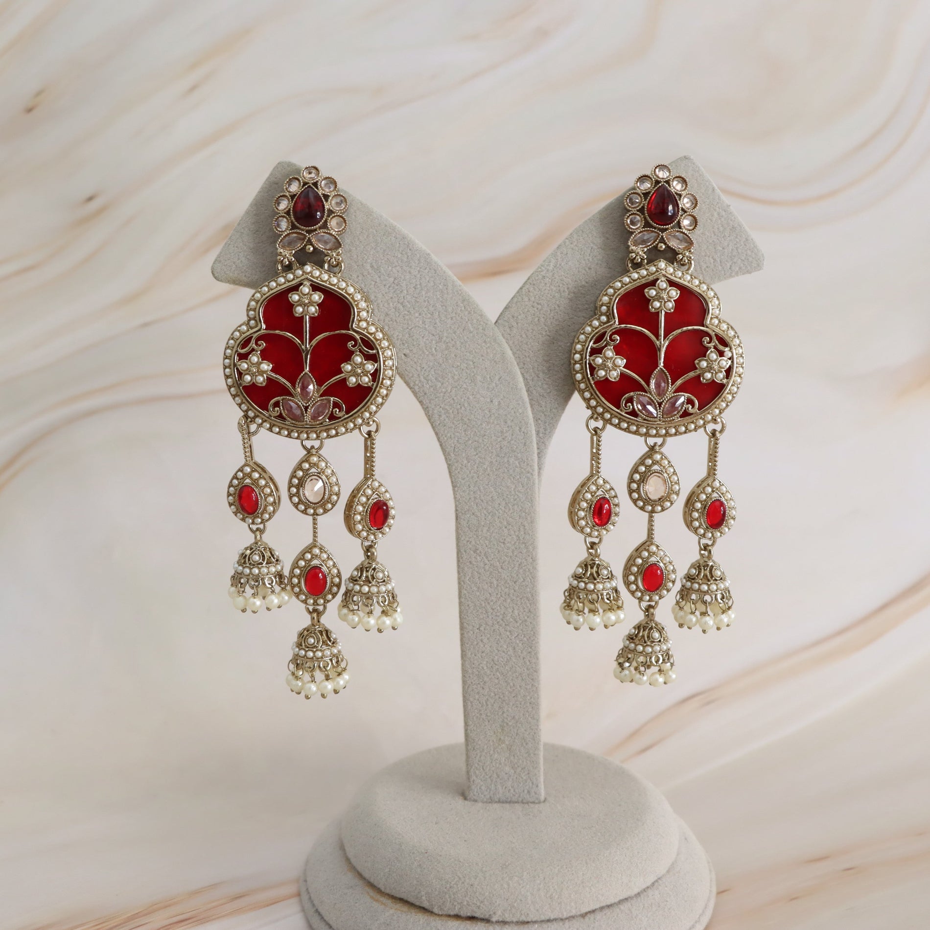 A pair of ornate Indian chandbali earrings with red enamel, gold-plated metalwork, and white faux pearls, displayed on a light gray jewelry stand against a marble-patterned background.