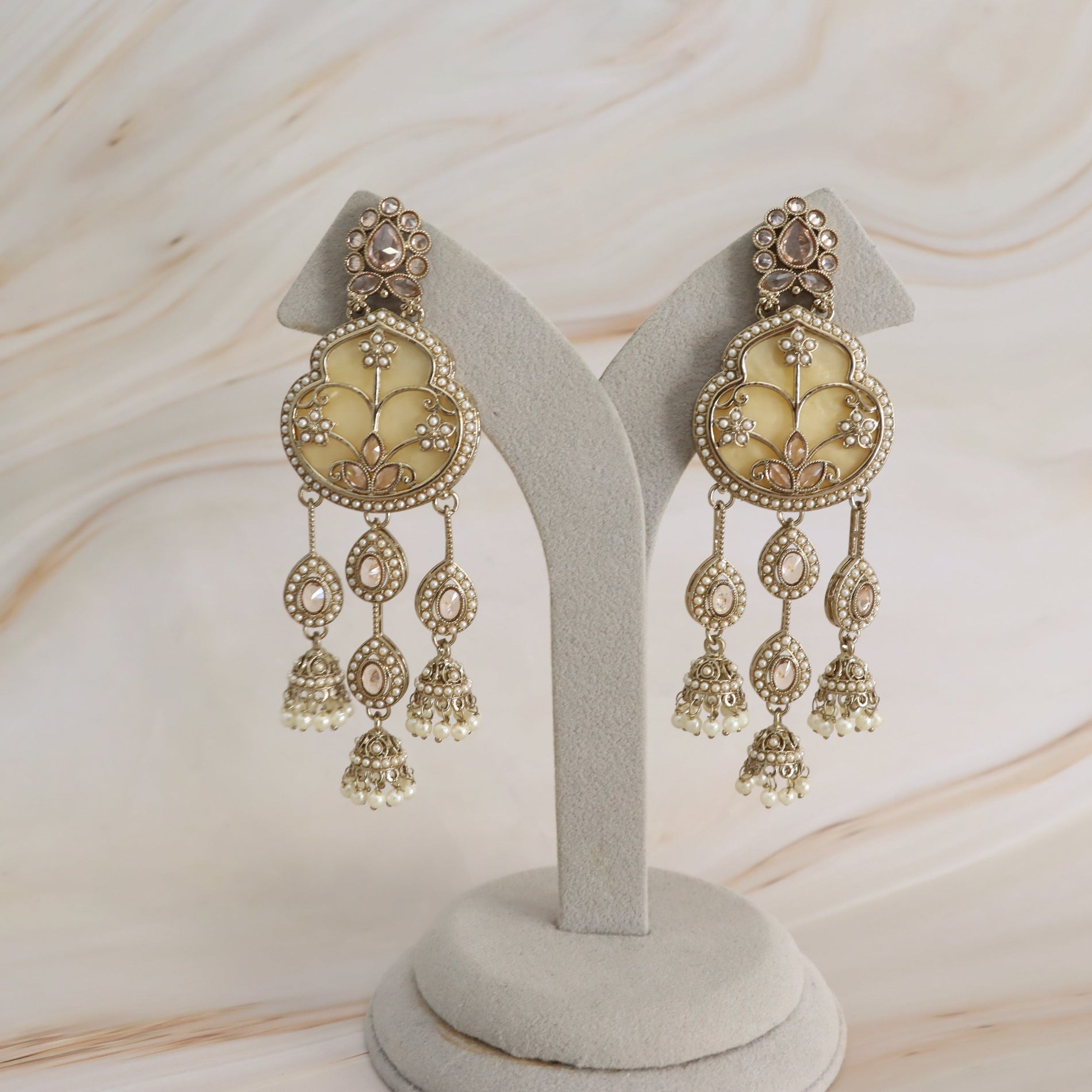 A pair of ornate gold-toned dangle earrings with cream enamel panels and small clear stones, displayed on a light gray jewelry stand against a soft beige marble-like background.