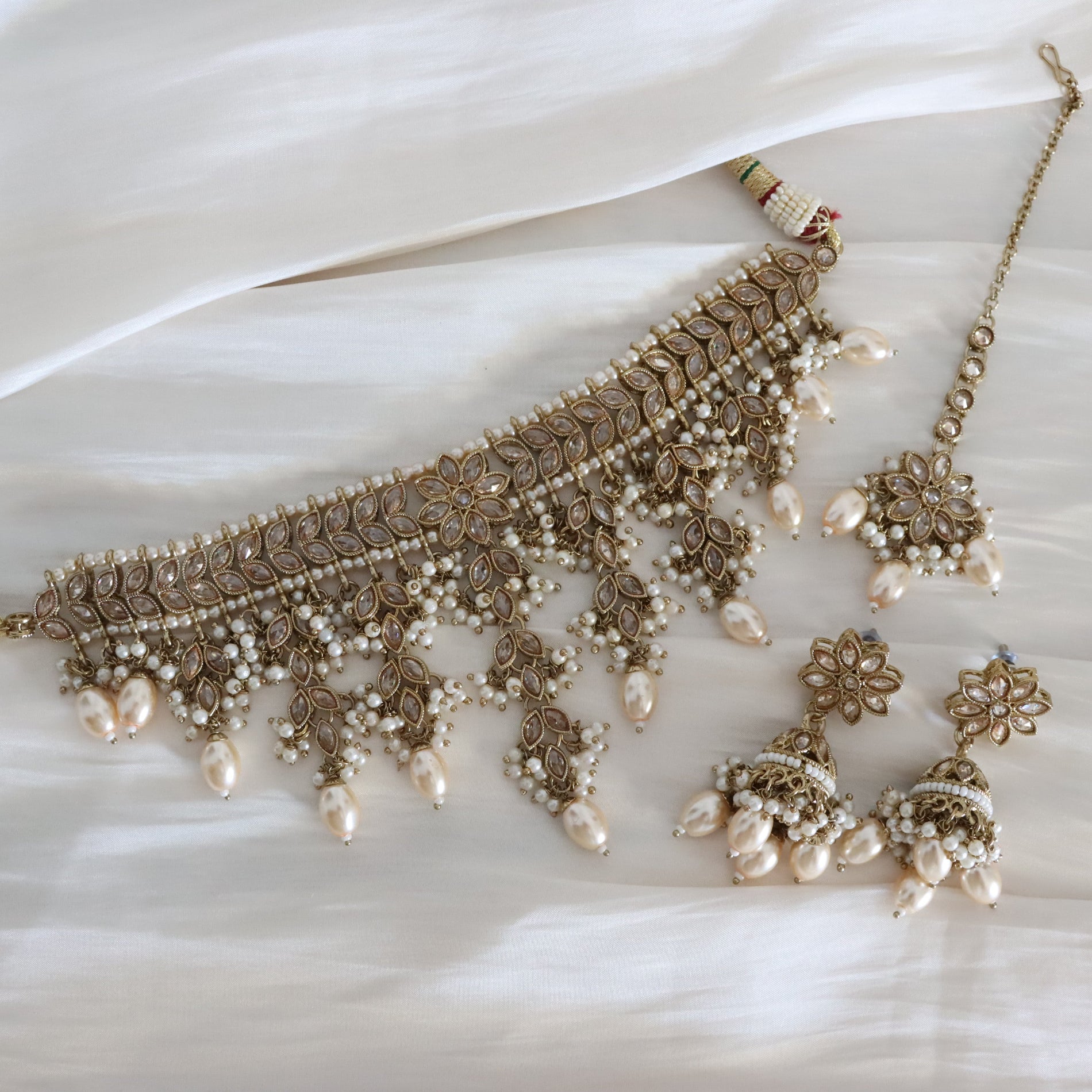 An ornate traditional Indian jewelry set featuring a gold-plated filigree necklace with dangling faux pearls, matching earrings, and a maang tikka, arranged on a draped white fabric background.