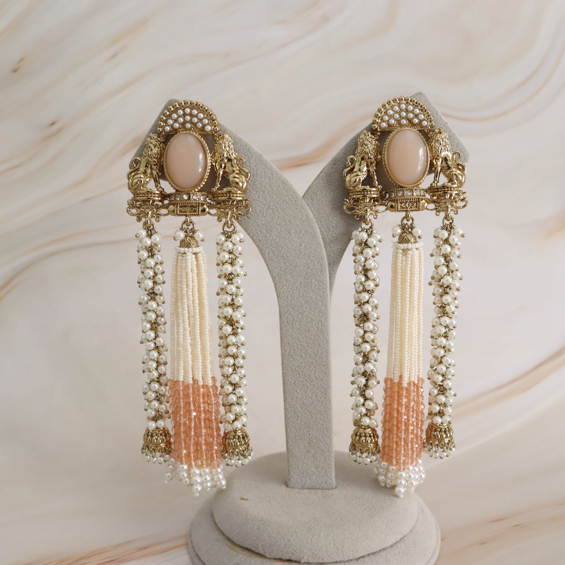 A pair of ornate gold-toned statement earrings displayed on a gray jewelry stand against a light marble-patterned background. Each earring features a peach-colored oval stone, intricate filigree detailing, and long dangling strands of cream beads and peach beaded tassels.