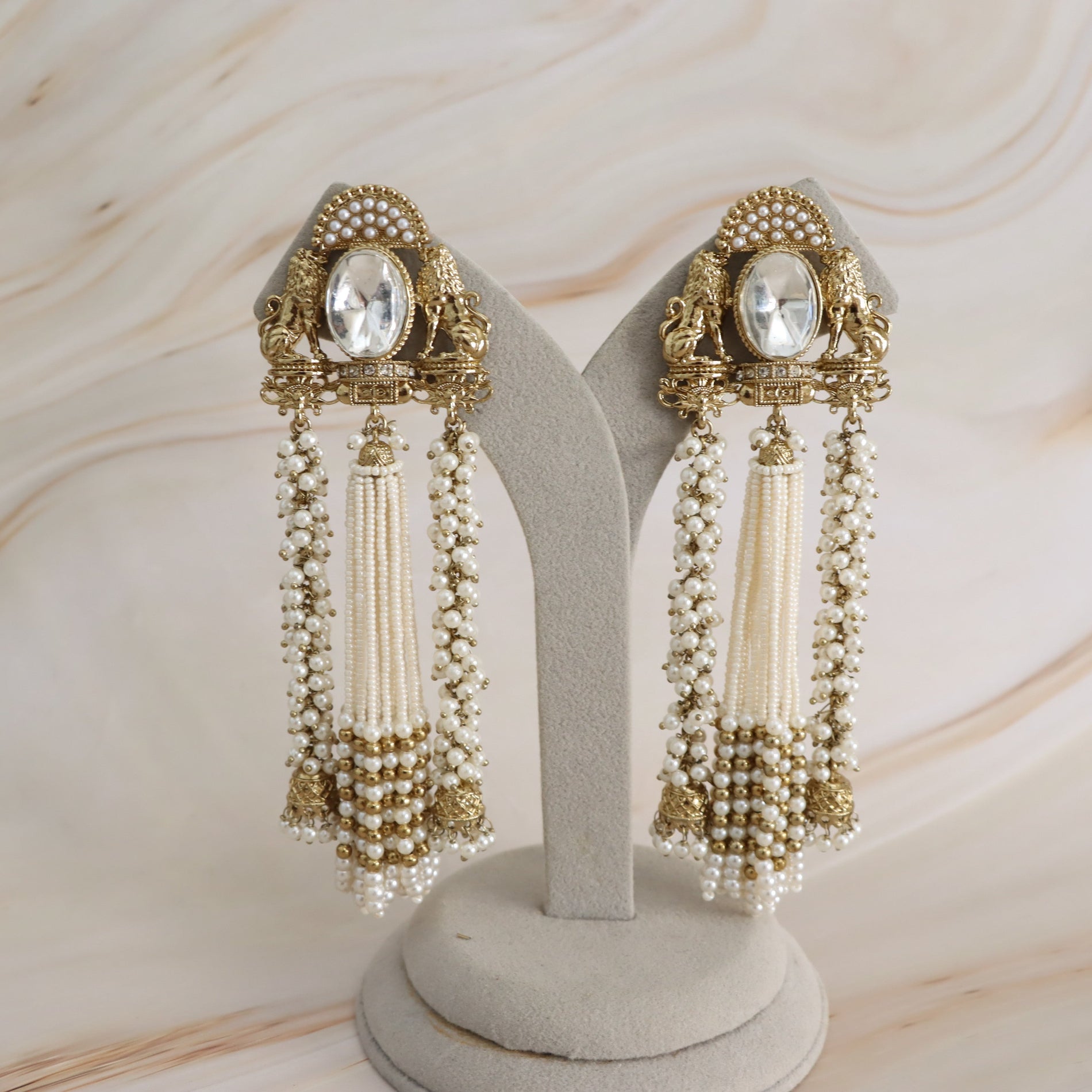 A pair of ornate gold and cream beaded dangle earrings displayed on a light gray jewelry stand against a soft beige marble background.