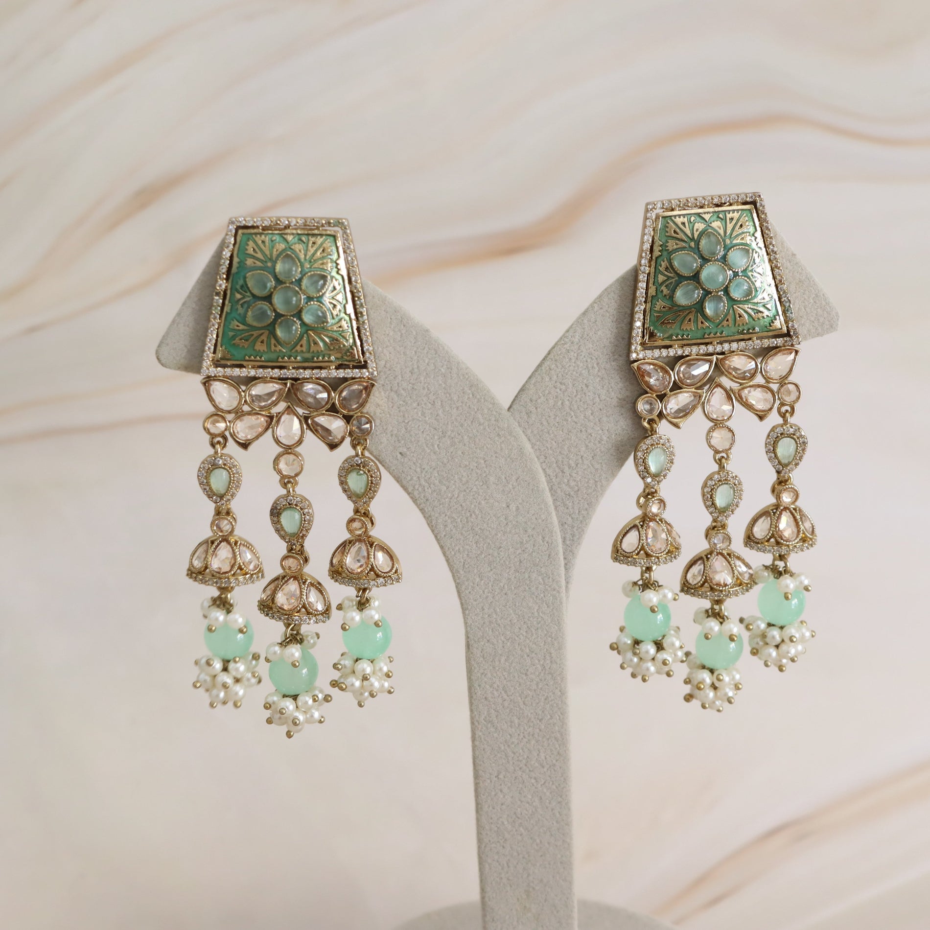 A pair of ornate gold-toned Indian jhumka earrings with green enamel floral motifs, hanging light blue stones, and white pearl-like accents, displayed on a gray jewelry stand against a light beige marble background.