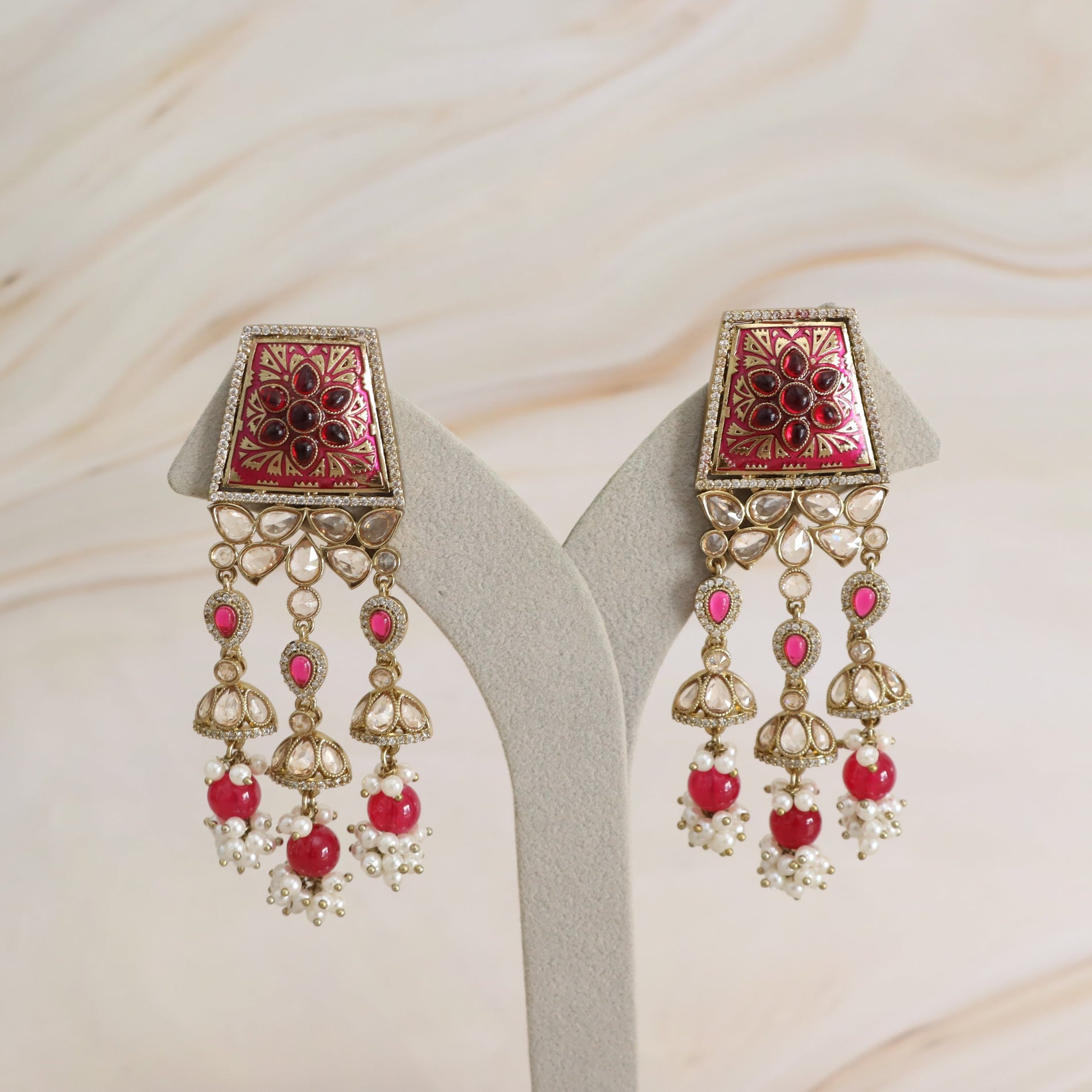 A pair of ornate gold-toned dangle earrings with pink floral enamel tops and red heart-shaped beads, displayed on a light gray jewelry stand against a soft beige marble background.