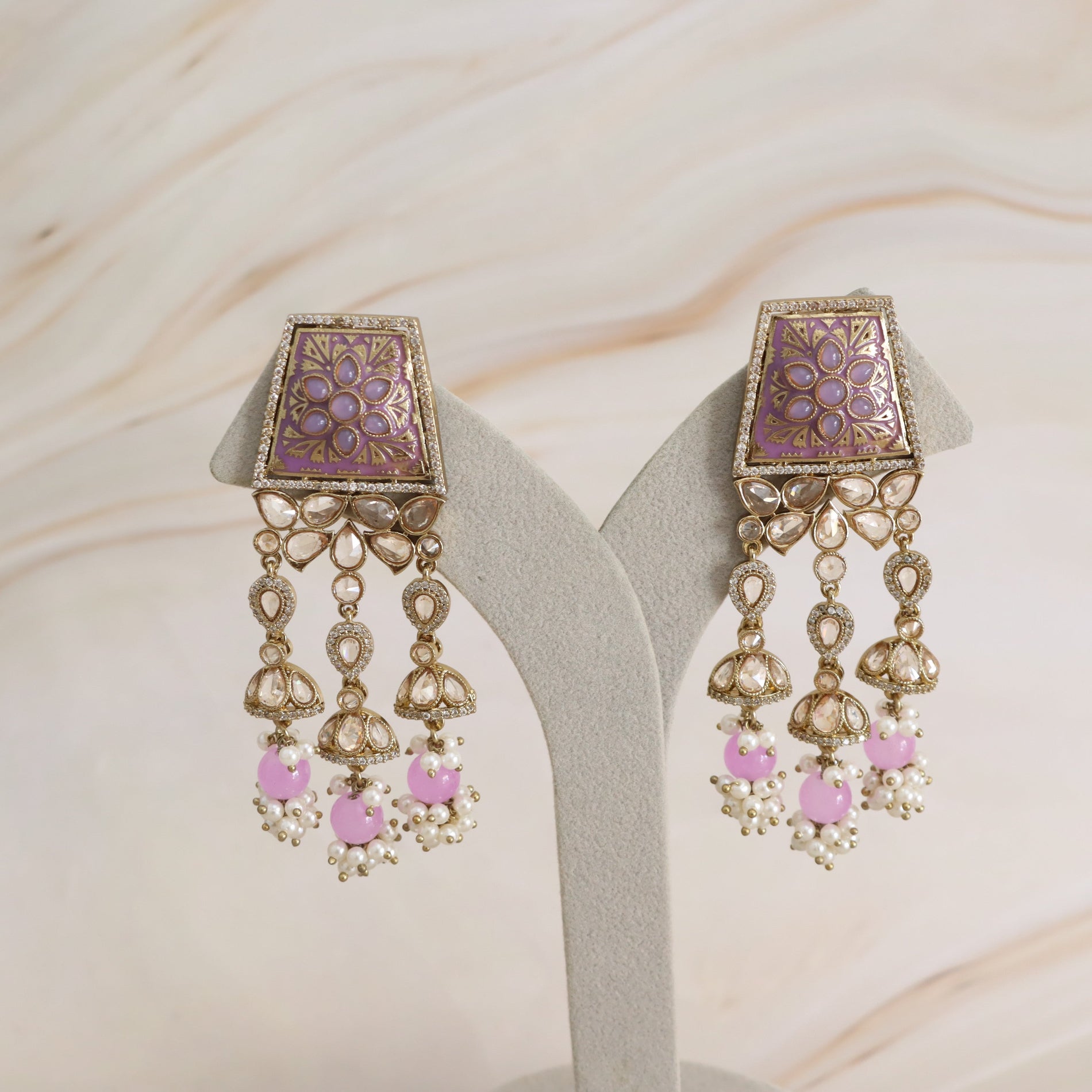 A pair of ornate gold-toned statement earrings displayed on a light gray jewelry stand against a soft beige marble background. The earrings feature square purple enamel panels with floral detailing, dangling gold-toned filigree, and pink heart-shaped beads accented with small white pearls.